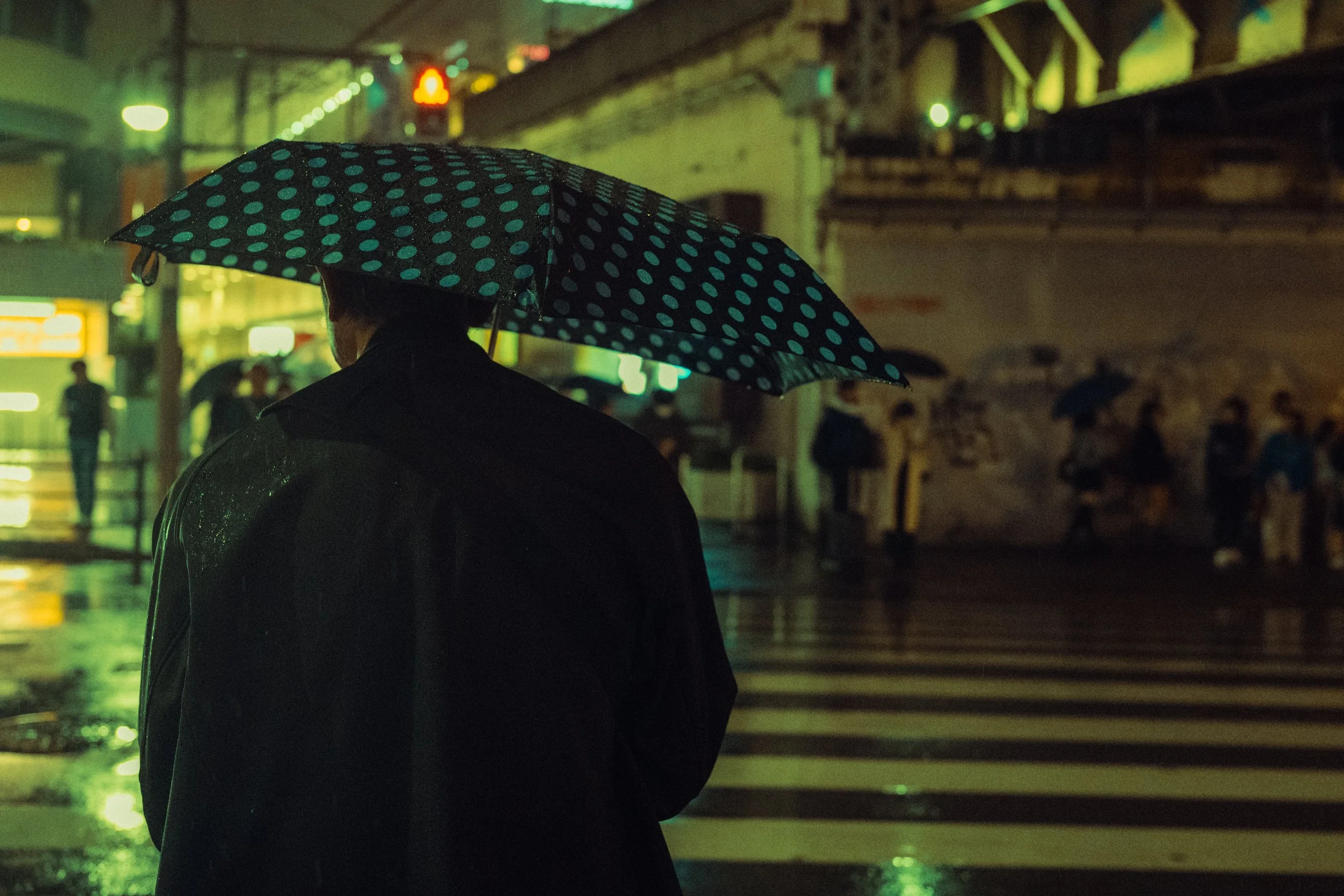 Personne avec un parapluie à pois bleus et blancs, debout dans la pluie la nuit, dans un environnement urbain éclairé.