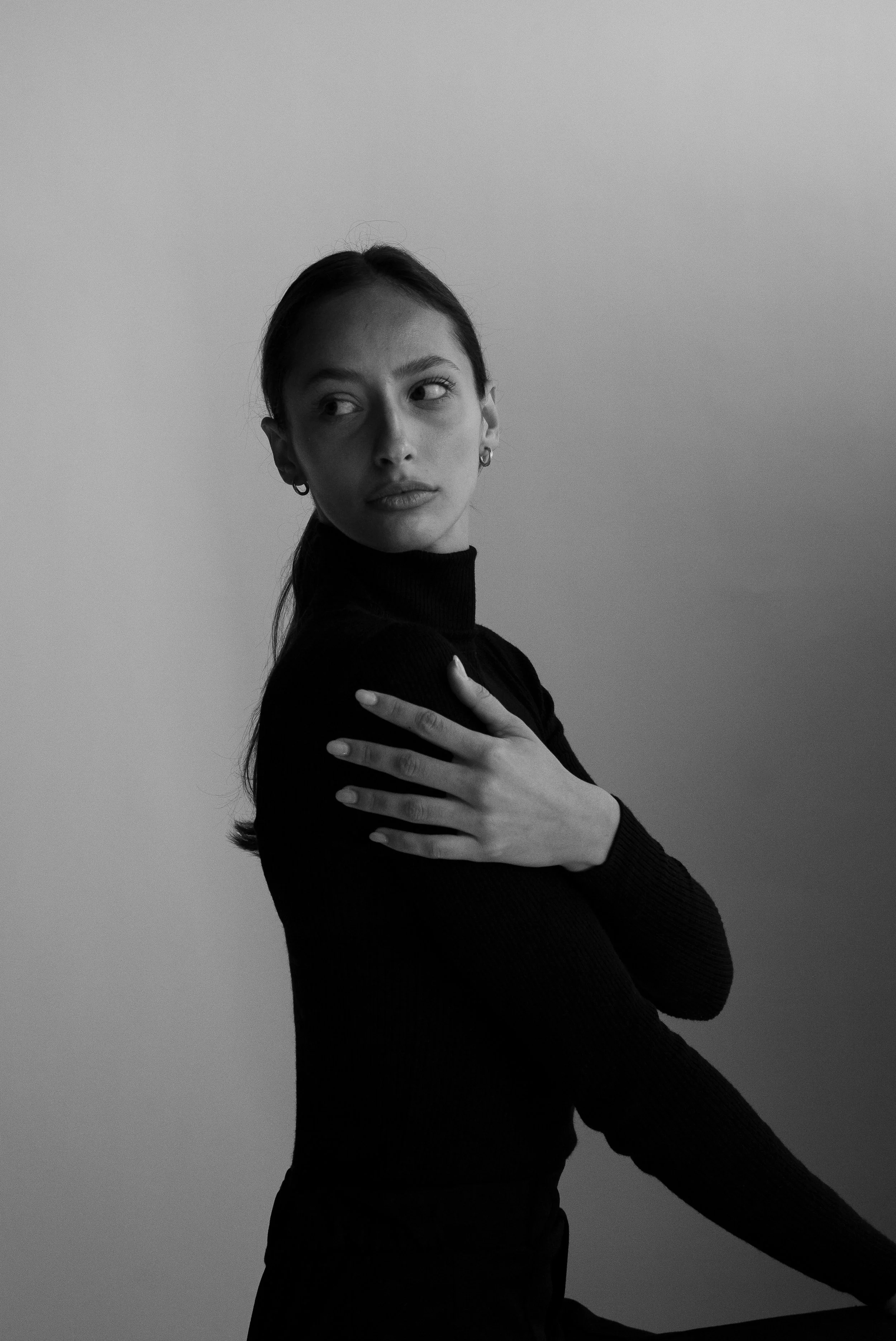 Une femme en noir et blanc avec les cheveux attachés, portant un pull à col roulé, regarde vers la gauche avec une expression calme, les bras croisés sur la poitrine.