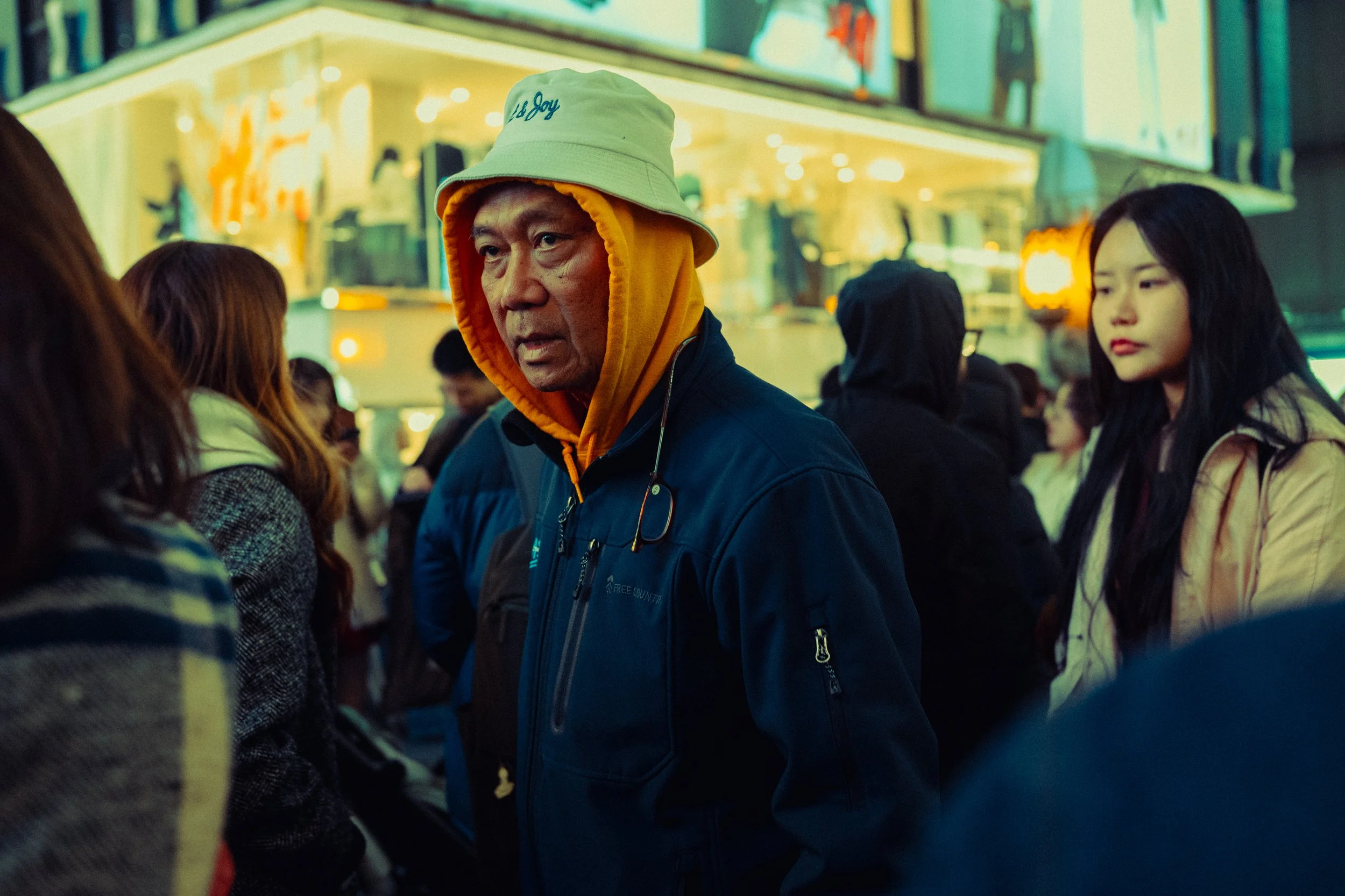Une personne portant une casquette beige, une veste bleue et un hoodie orange dans une foule urbaine en soirée.
