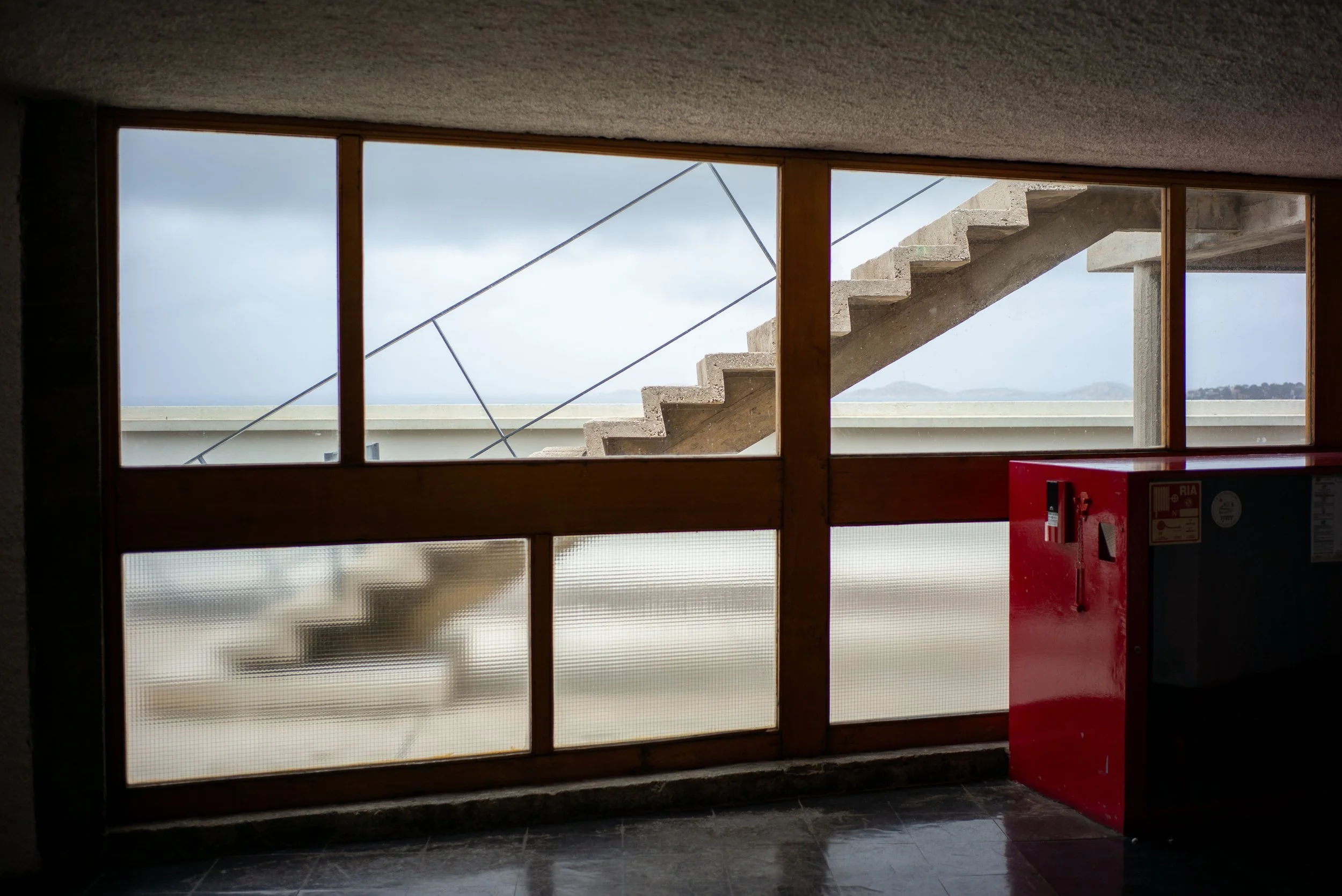 Vue à travers une fenêtre en bois avec un cadre en acier, montrant un escalier en béton à l'extérieur, un ciel nuageux et des montagnes au loin. Il y a aussi un coffre rouge dans l'intérieur.