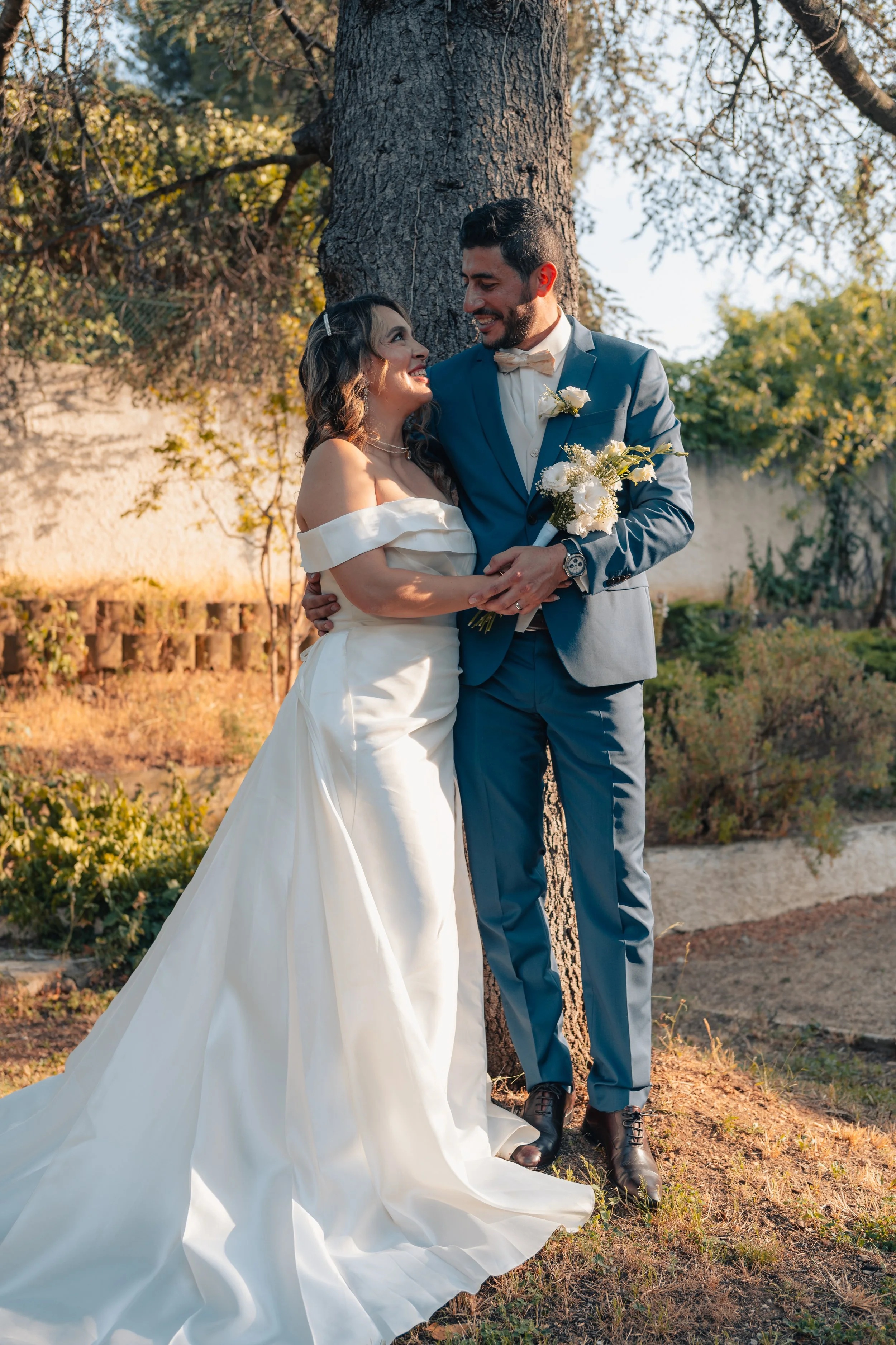 Un couple en tenue de mariage, souriant et regardant l'un l'autre, à l'extérieur près d'un arbre, avec un décor naturel et ensoleillé.