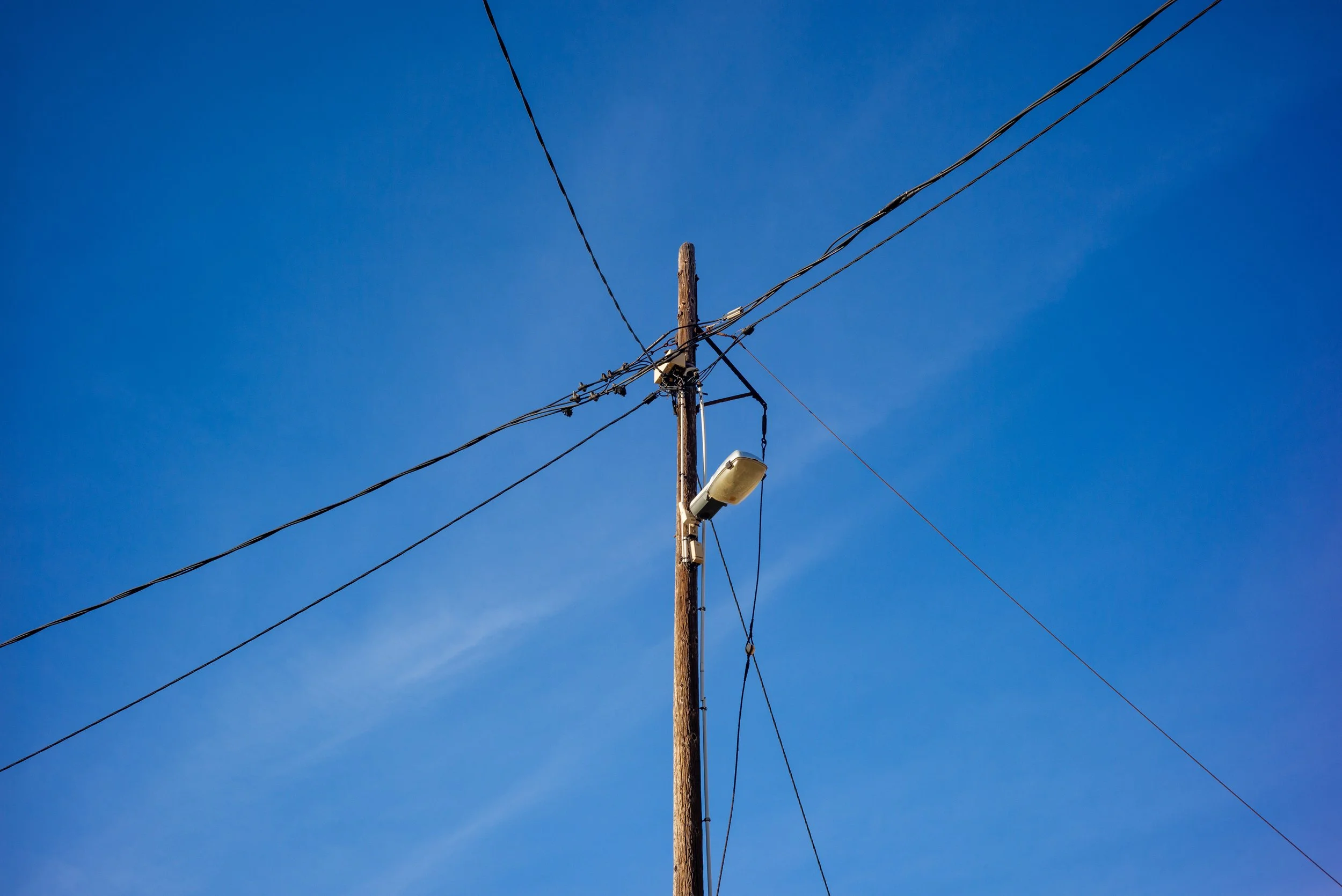 Poteau électrique en bois avec un lampadaire fixé et plusieurs câbles électriques contre un ciel bleu clair.