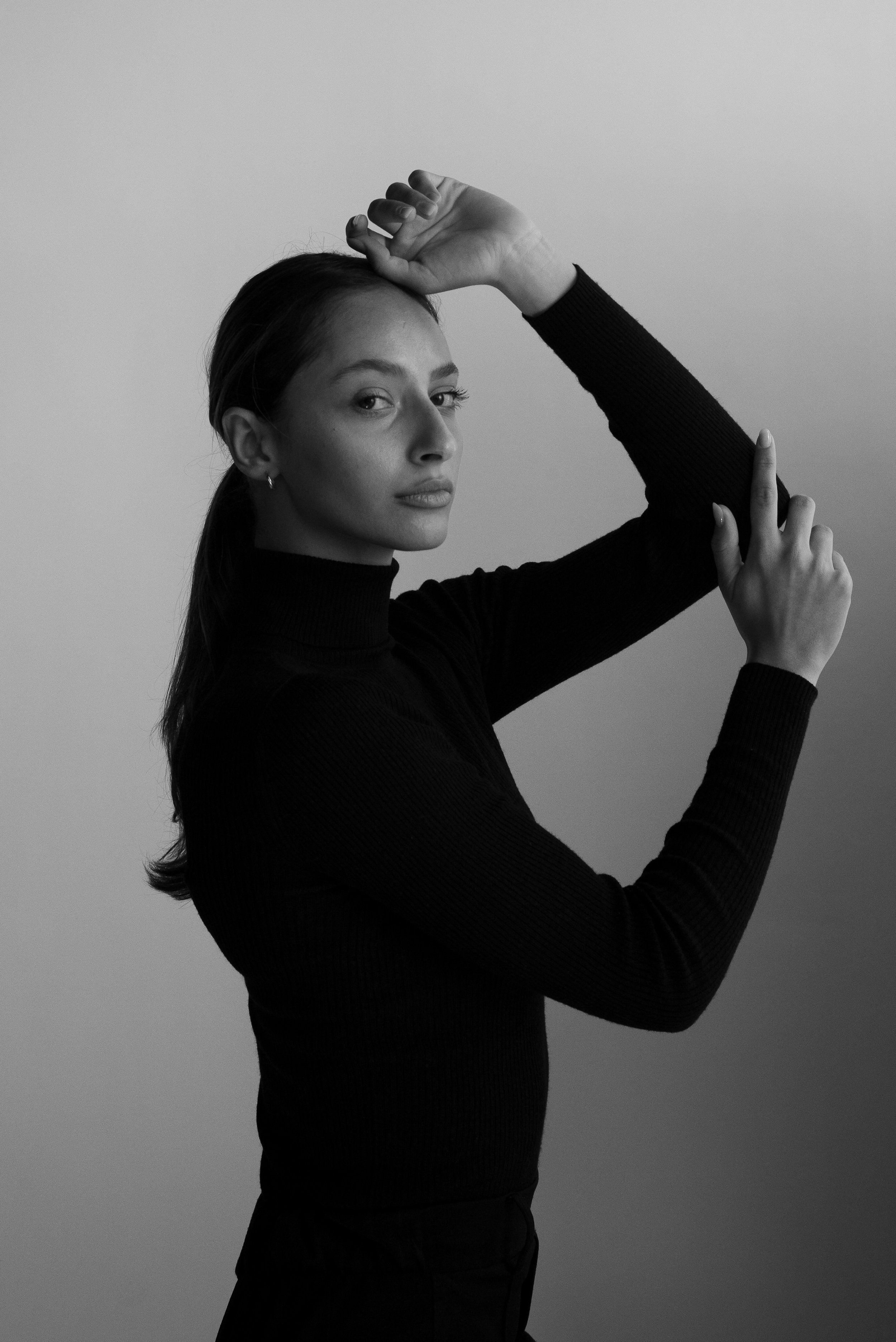 Portrait en noir et blanc d'une femme avec les bras levés, portant un pull à col roulé.