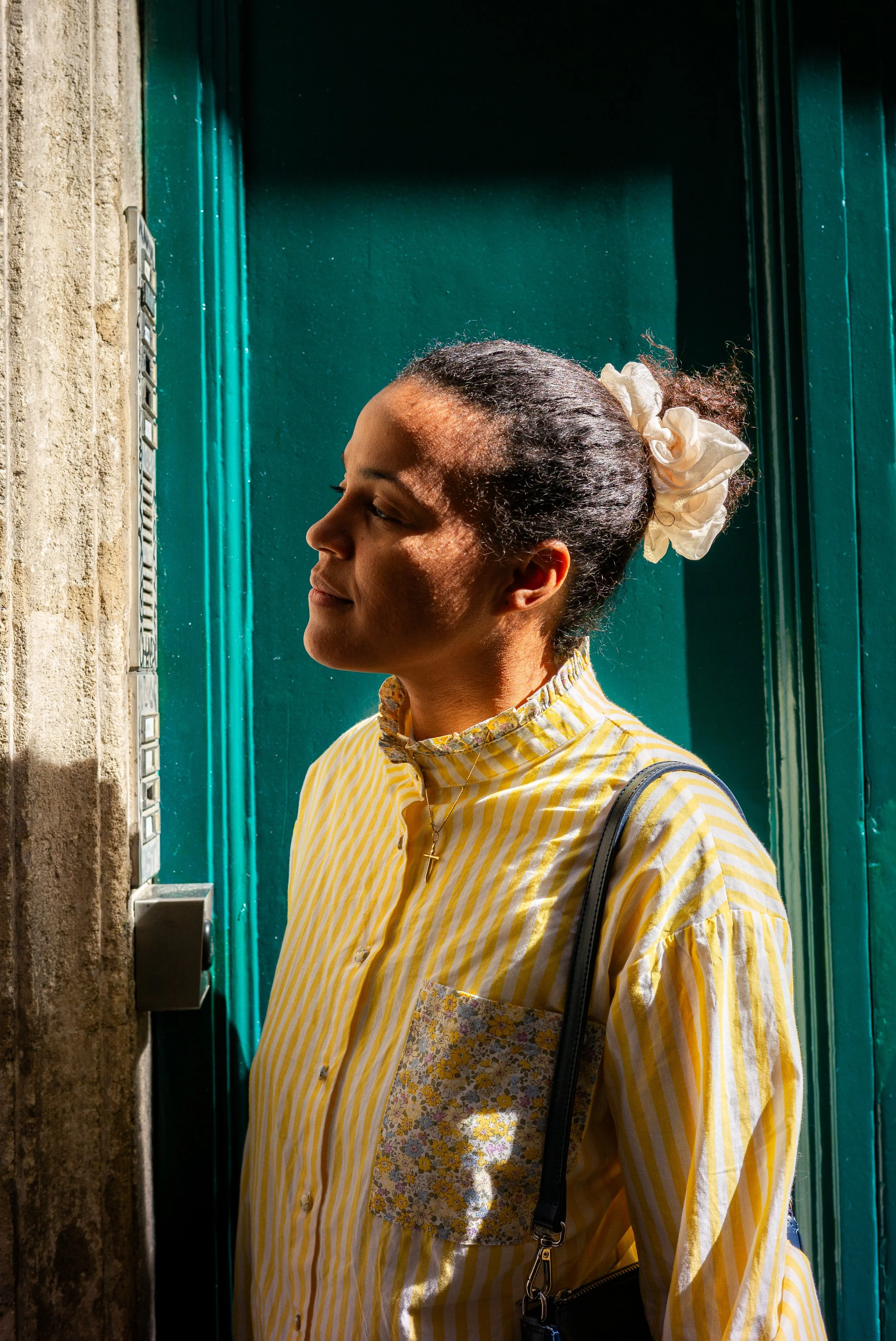 Une femme souriante portant une chemise jaune à rayures, regardant un interphone avec une expression de plaisir ou de curiosité, avec un pavé en pierre sur le côté gauche et une porte verte en arrière-plan.
