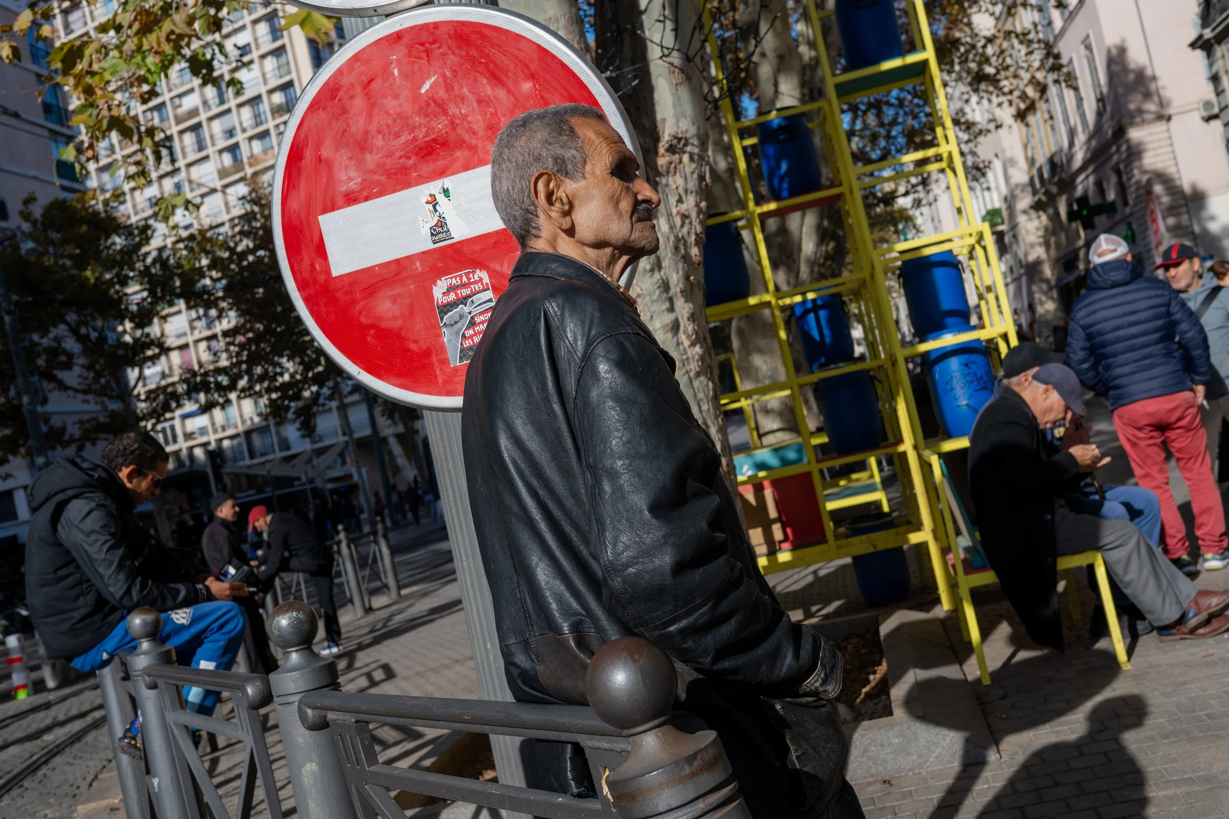 Un homme en cuir noir assis contre une barrière en métal sur un trottoir urbain, avec un panneau d'interdiction derrière lui, d'autres personnes assises ou debout, et une structure jaune avec des barils bleus à l'arrière.