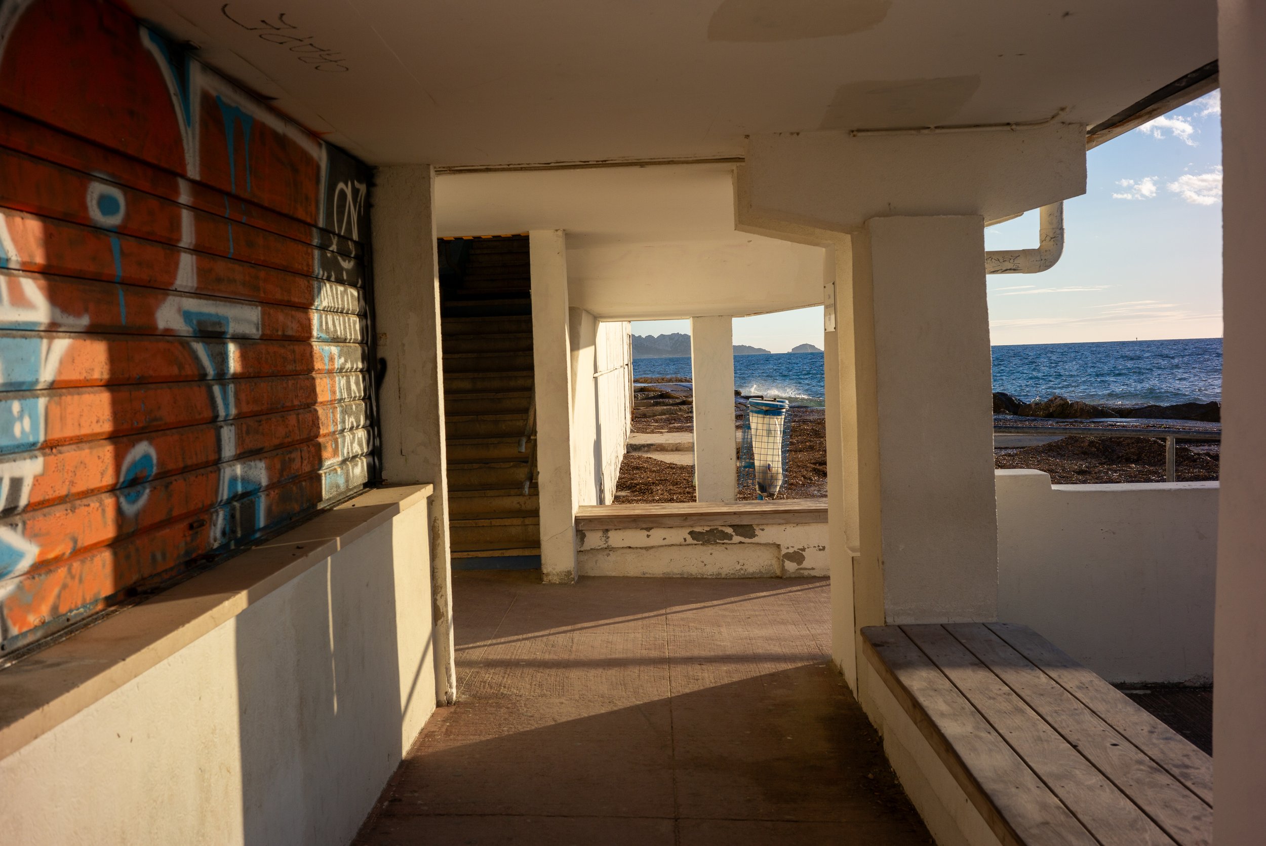 Vue intérieure d'une structure en béton donnant sur une plage avec vue sur la mer, un bac à poubelles bleu et une escaliers, avec un ciel partiellement nuageux.