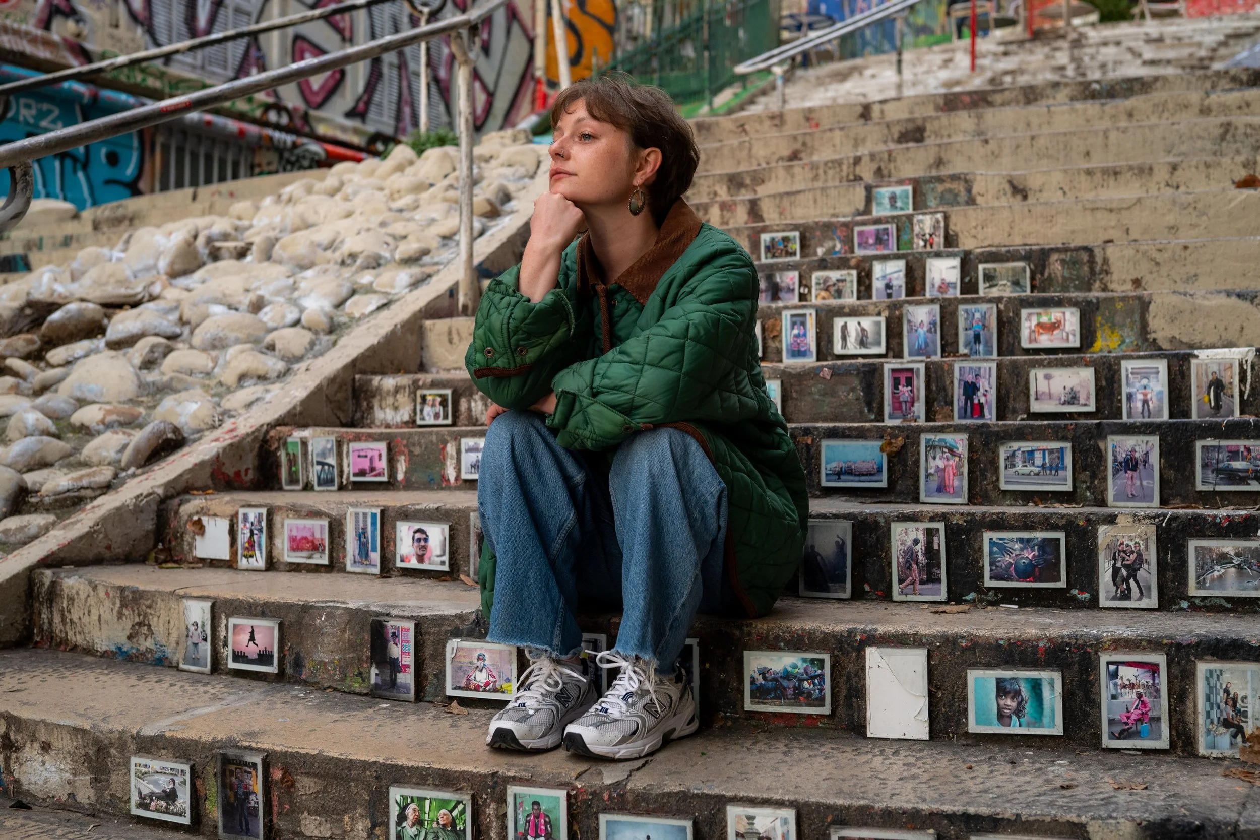 Une femme assise sur des escaliers, entourée de nombreuses photographies exposées sur les marches, dans un environnement urbain avec des graffitis en arrière-plan.