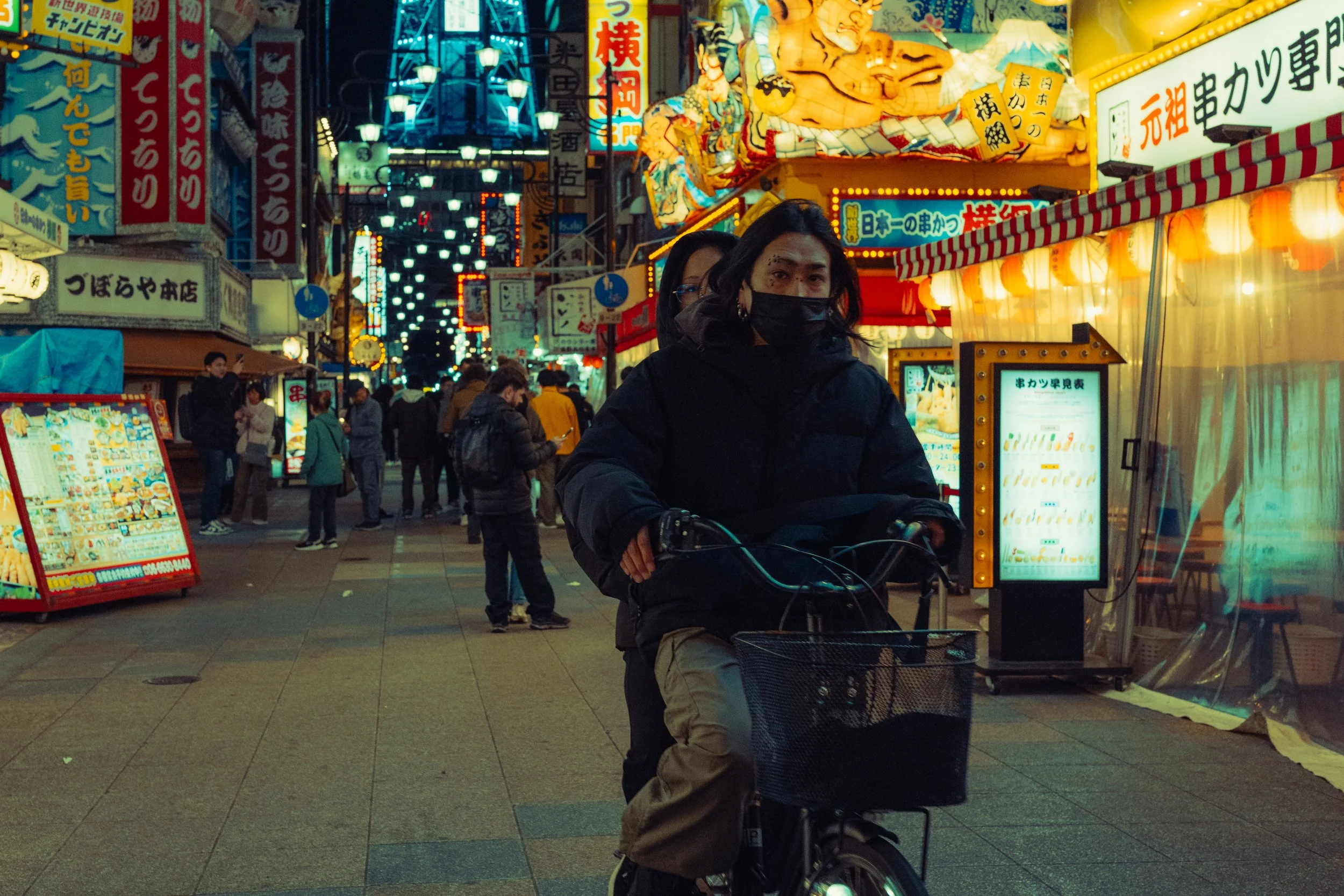 Une personne à vélo portant un masque noir dans une rue animée éclairée de néons, avec plusieurs autres personnes et des enseignes en japonais.