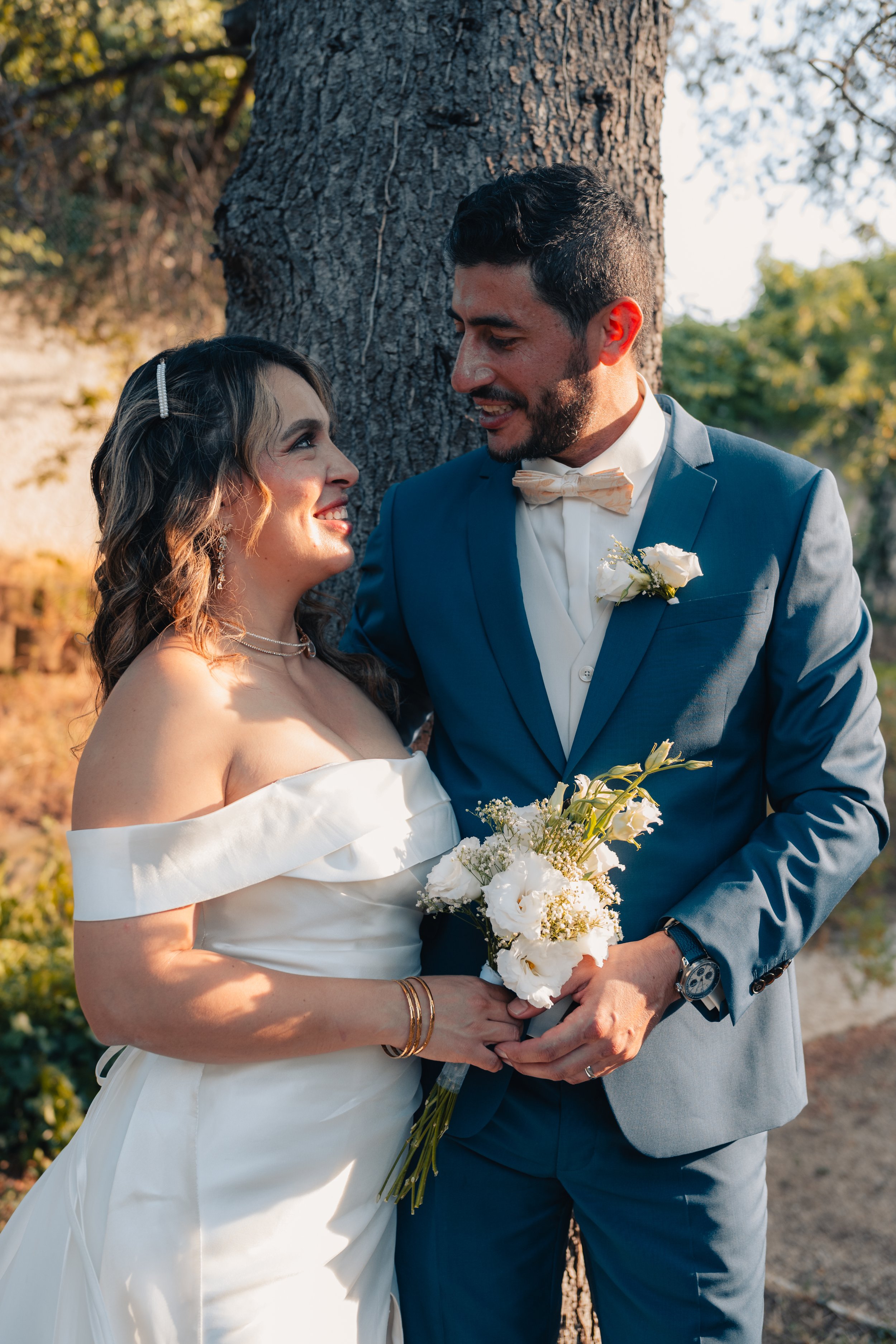 Un couple en costume de mariage se regarde tendrement devant un arbre en plein air, la femme tient un bouquet de fleurs blanches.