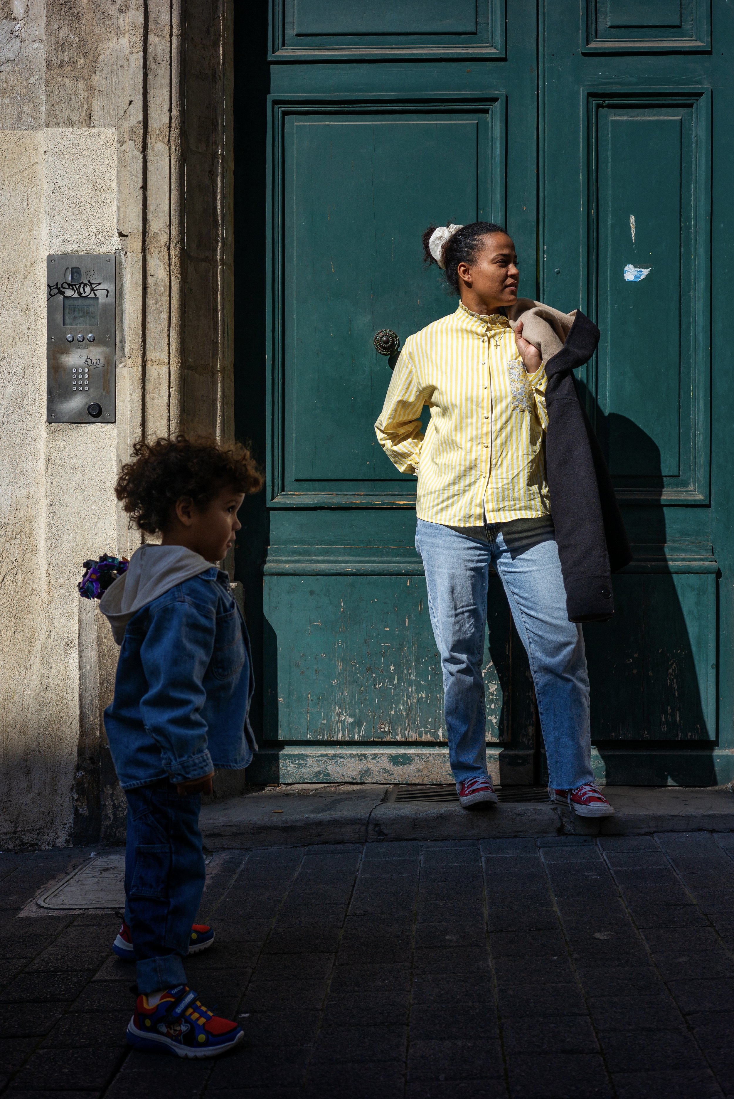 Une femme et un jeune garçon debout devant une grande porte verte. La femme porte une chemise jaune à rayures et tient un manteau noir. Le garçon porte un jean et une veste en denim. Le sol est pavé et un interphone est fixé sur le mur à gauche.