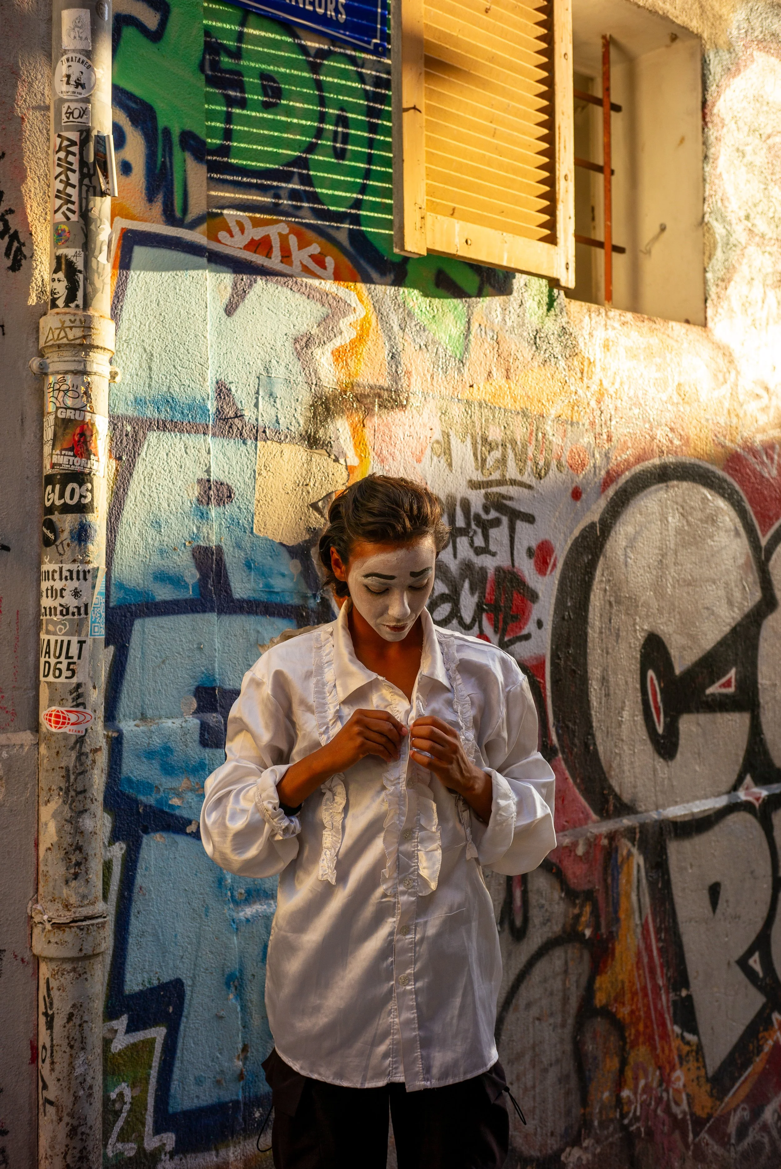 Une jeune femme debout contre un mur graffité, portant une chemise blanche et ajustant ses vêtements, avec un œil maquillé en noir et un fond coloré.