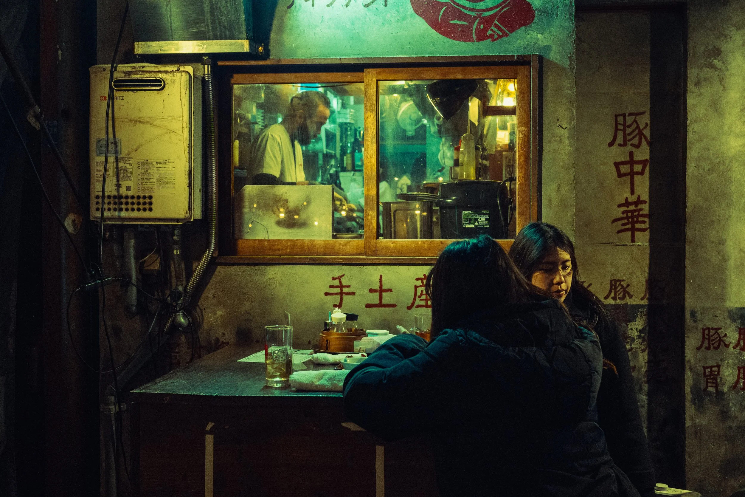 Deux femmes assises à une table dans un restaurant asiatique, avec un chef préparant des plats derrière une fenêtre en bois, dans un décor aux murs en béton avec des caractères chinois rouges.