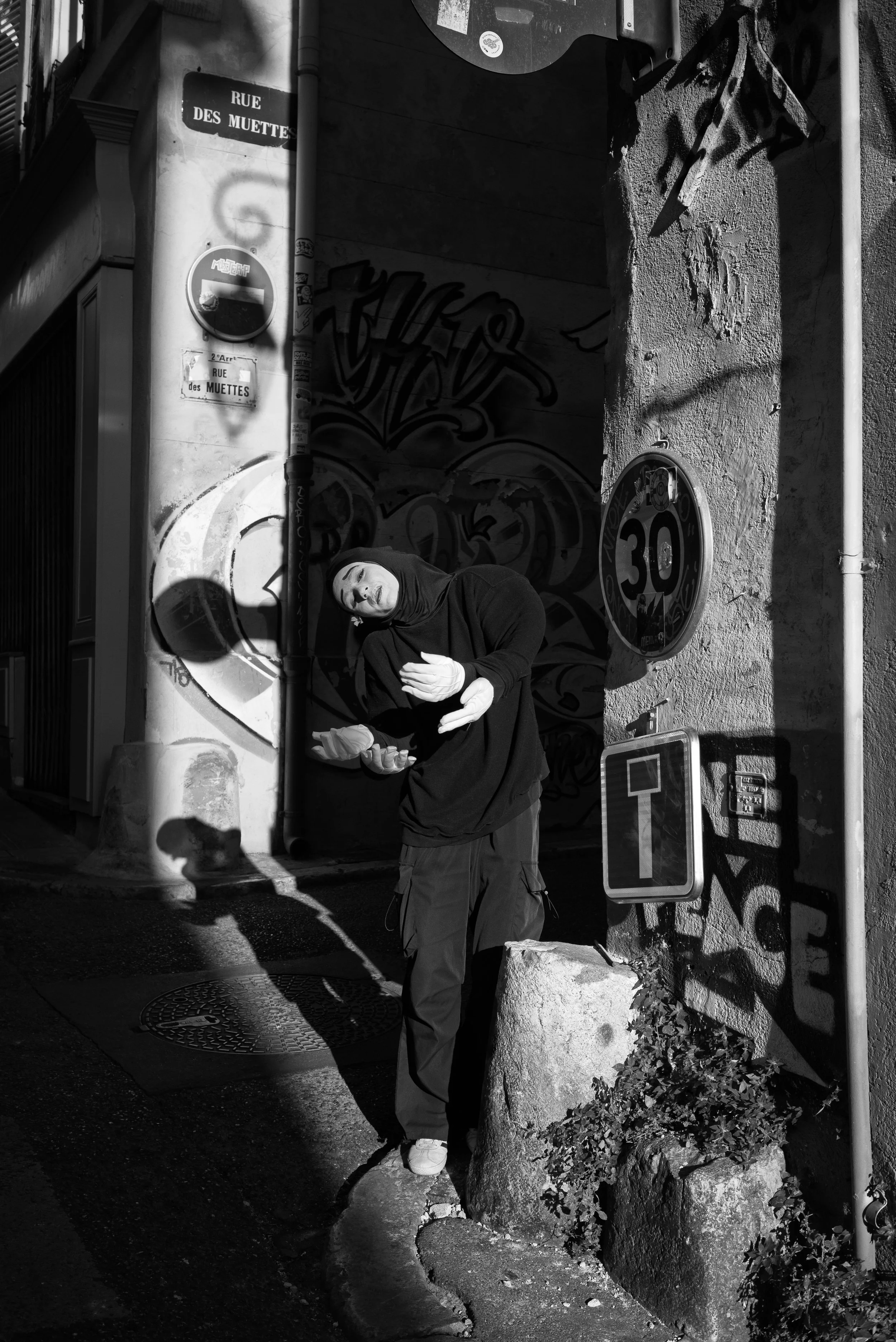 Un homme portant un hoodie noir et un pantalon de cargo porte une paire de gants blancs, debout près d'un arbre en pierre, dans une rue pavée de Paris, avec des graffitis et des panneaux de signalisation