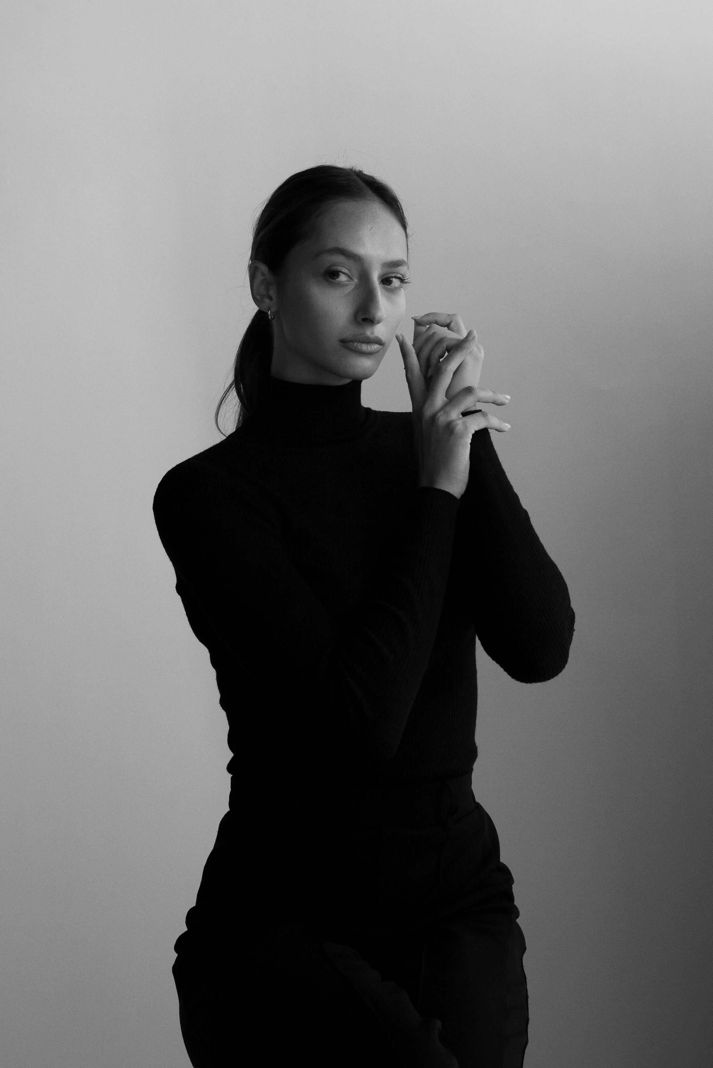 Portrait en noir et blanc d'une femme portant unpull noir, cheveux attachés, regardant vers l'objectif, posant avec ses mains près du visage contre un fond clair.