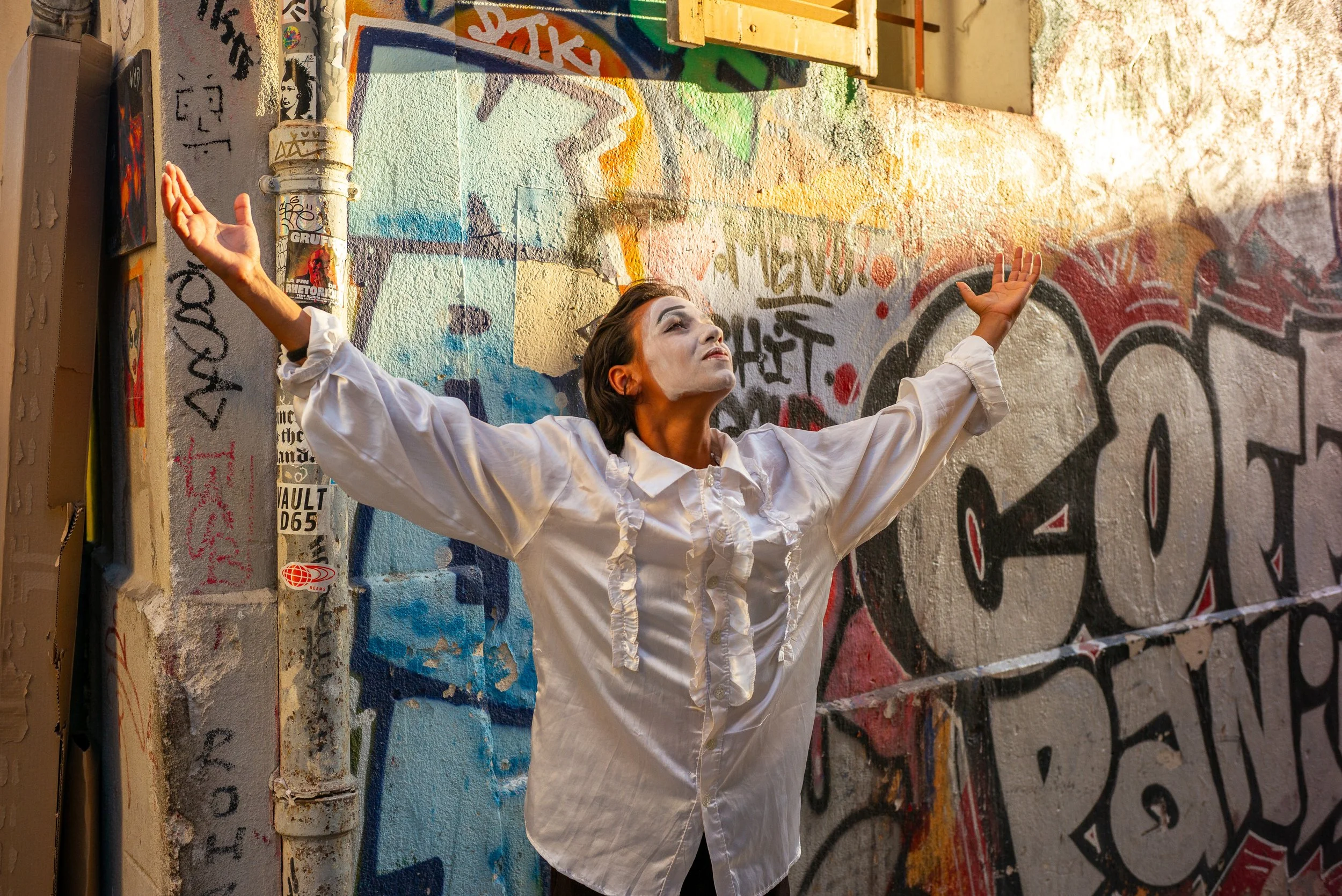 Une femme avec des vêtements blancs face à un mur couvert de graffitis, bras levés et regardant vers le haut, dans un environnement urbain