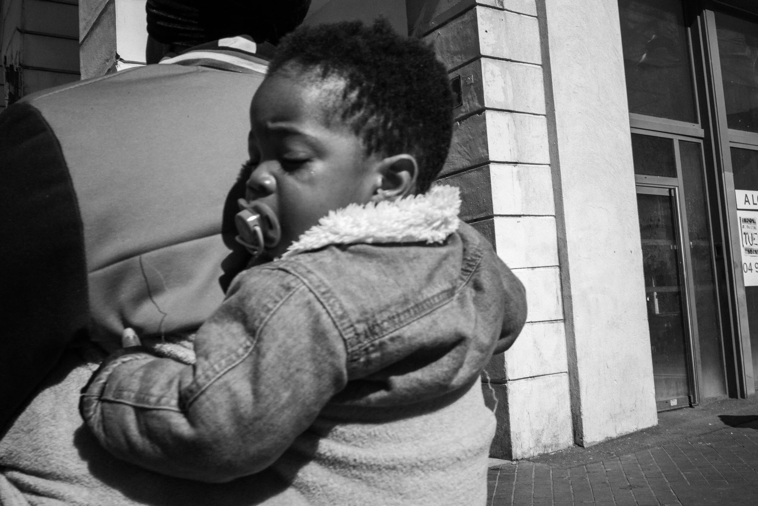 Un jeune enfant noir avec un bonnet en laine, utilisant une tétine, qui repose contre une personne plus âgée, devant un bâtiment en pierre.