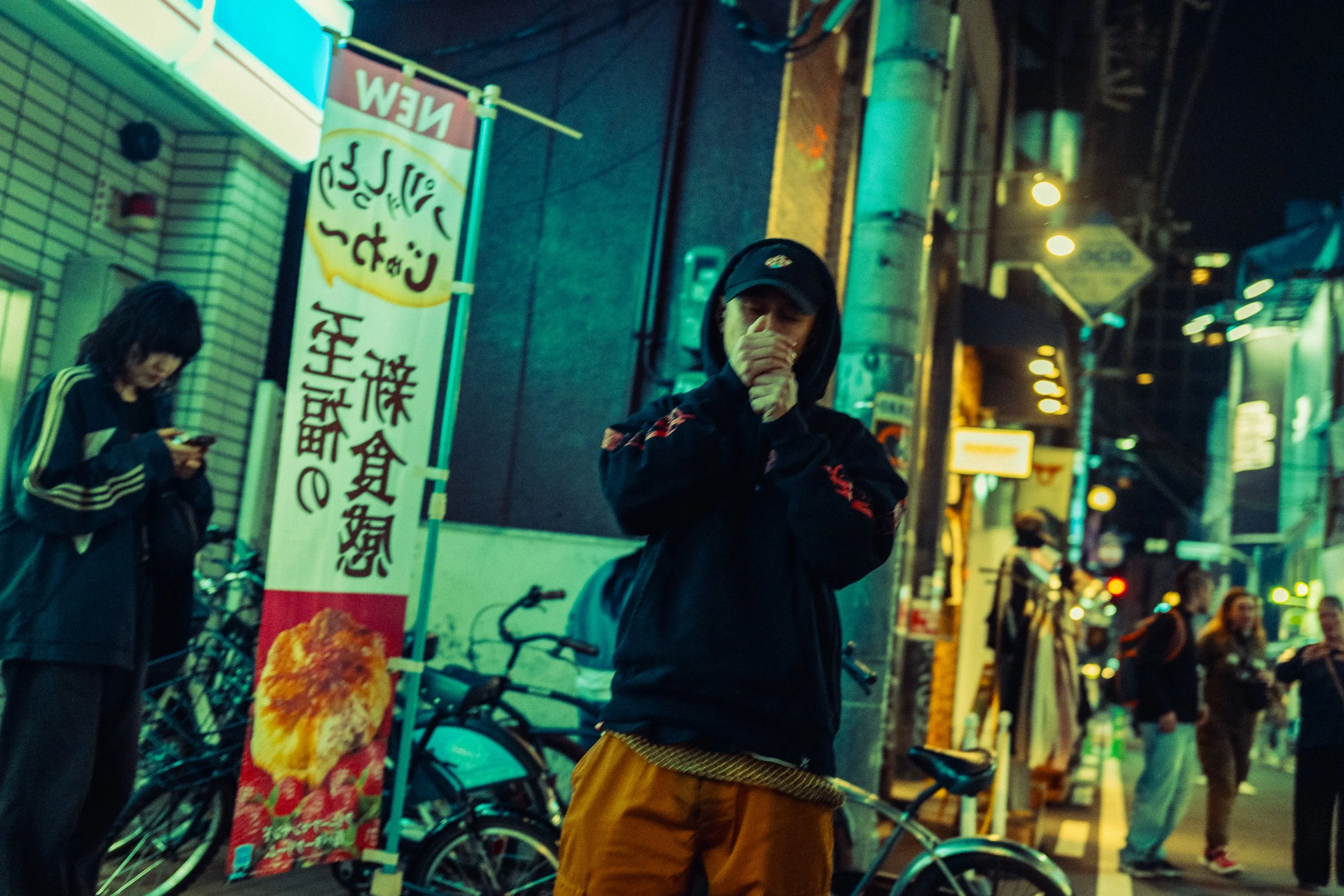 Une scène urbaine nocturne avec plusieurs personnes, notamment un homme en avant portant un hoodie noir et un masque, et une femme utilisant son téléphone à gauche. Il y a des vélos en arrière-plan, des enseignes lumineuses et des passants dans une r