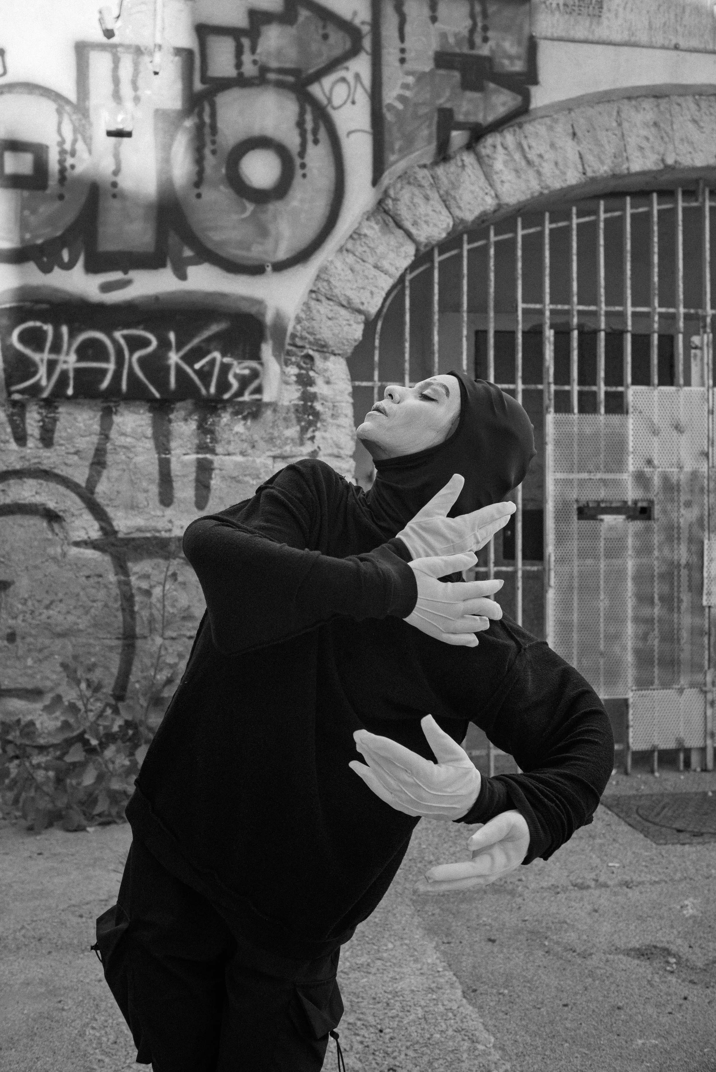 Une femme habillée en noir, portant des gants blancs, pose dans une ambiance urbaine avec des graffitis sur un mur en arrière-plan. Elle est en position de danse ou de performance artistique, avec une expression de concentration ou d'émotion.