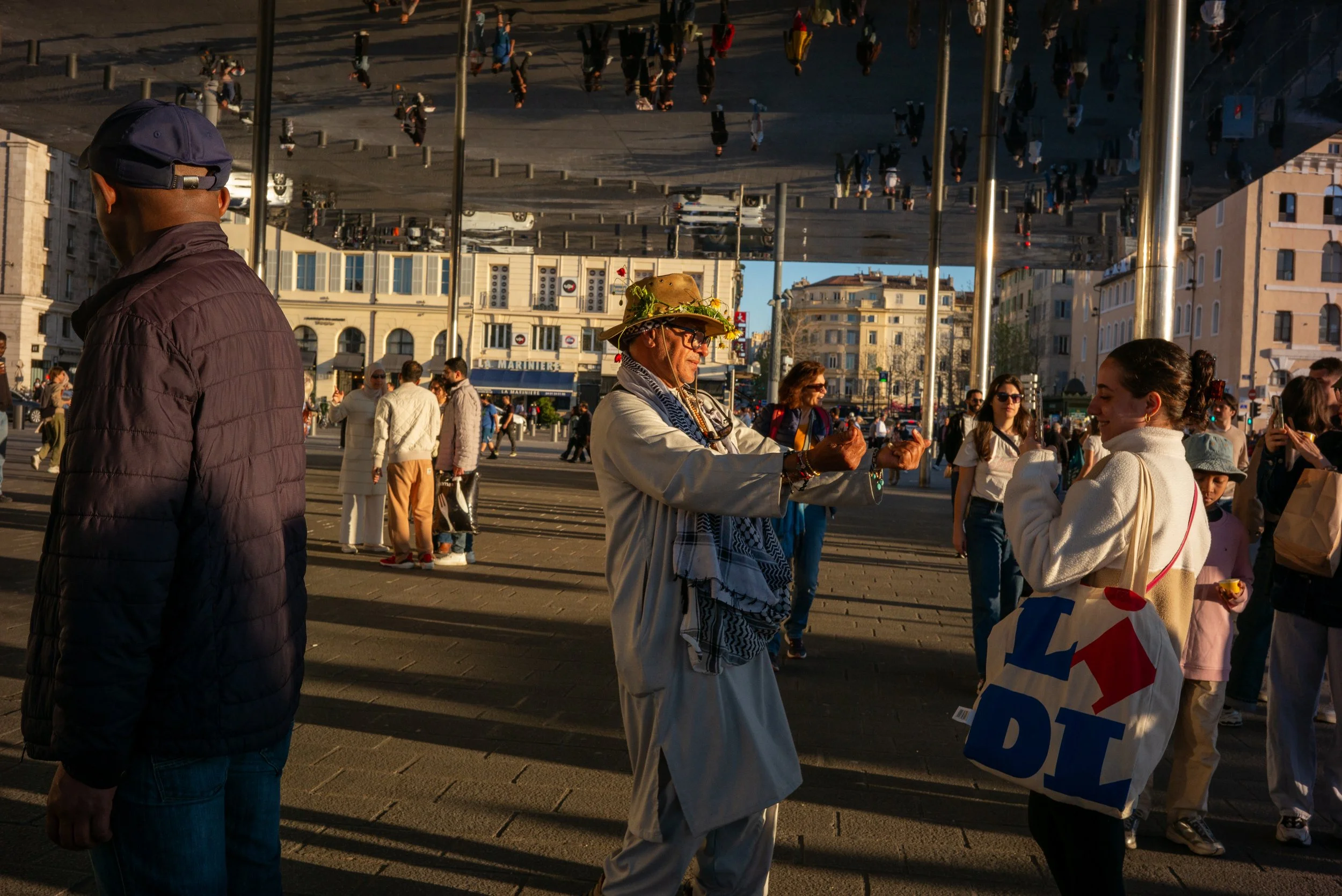 Un homme déguisé en moine avec un chapeau en fleurs fait un geste de la main vers une femme avec un sac à provisions blanc et un sac à main rouge, dans une rue animée avec plusieurs personnes et bâtiments en arrière-plan.