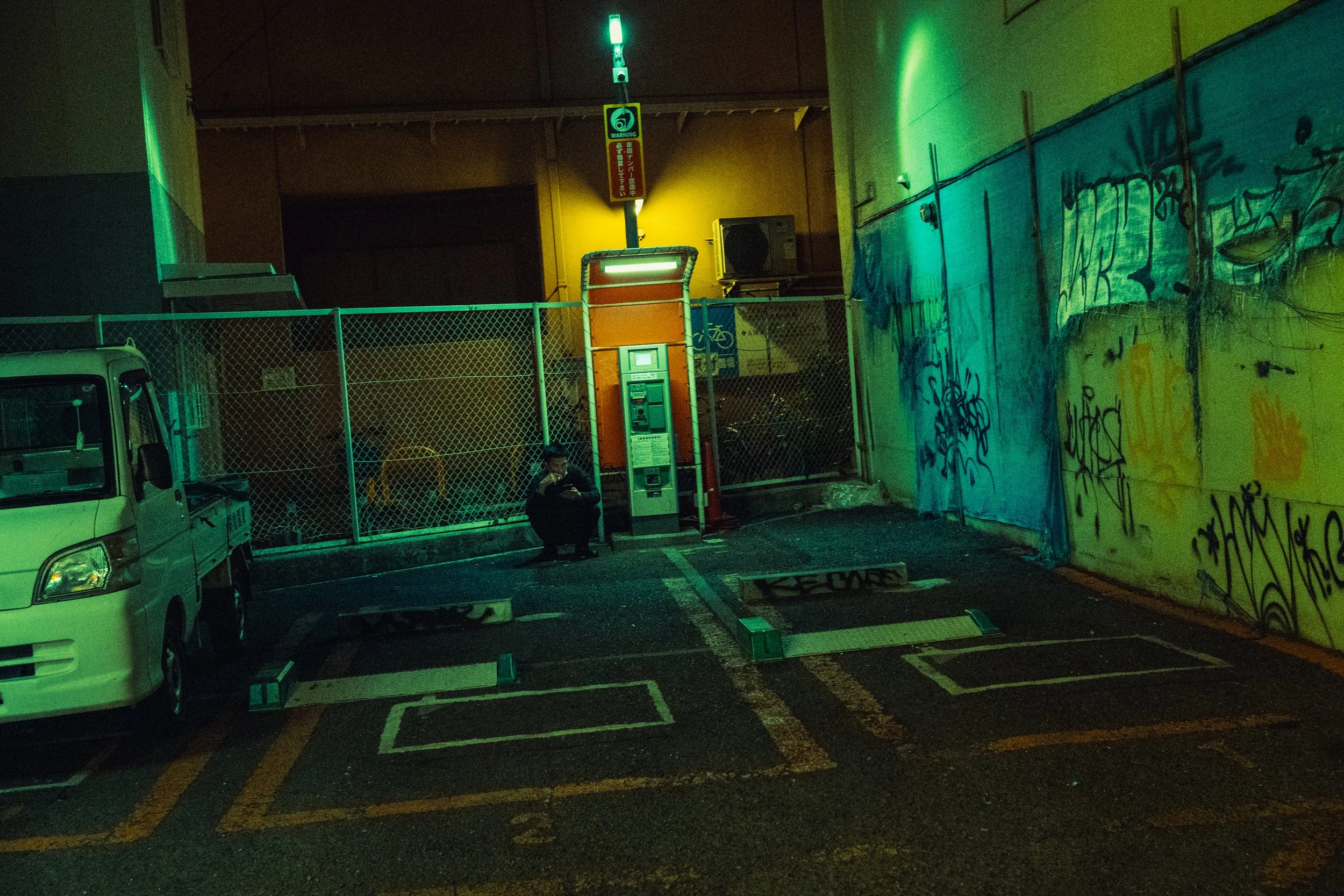 Un parking nocturne avec une personne assise près d'un distributeur de billets, entouré de graffitis sur un mur, une voiture blanche garée, et un panneau lumineux vert en haut.