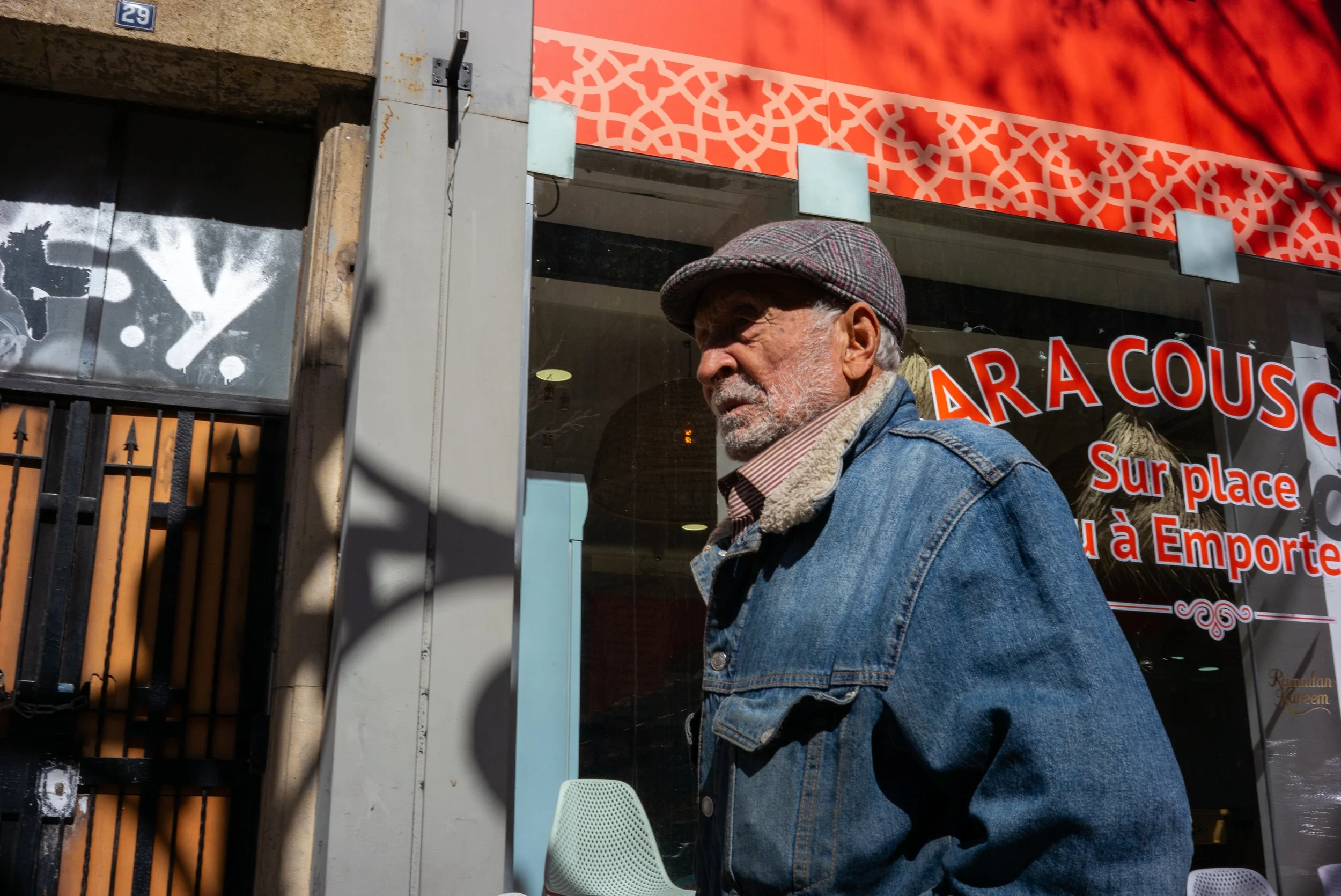 Un homme âgé porte une casquette à carreaux et un jean, se tenant près d'une vitrine de restaurant avec un panneau en arrière-plan.