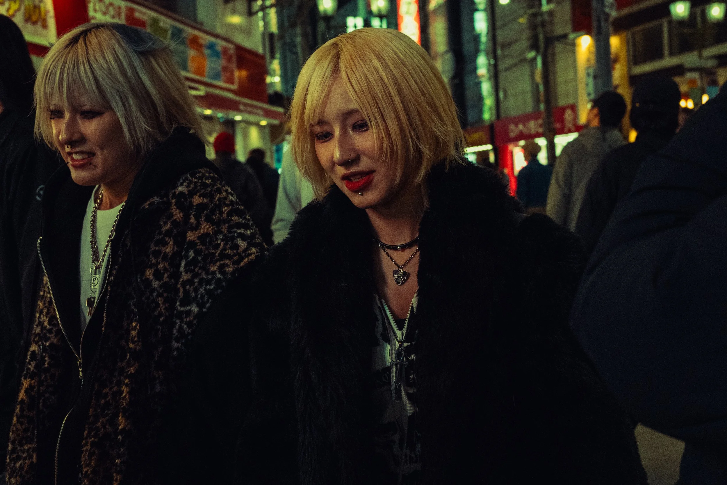 Deux jeunes femmes avec des cheveux blonds dans une rue urbaine la nuit, entourées de passants et de néons lumineux.