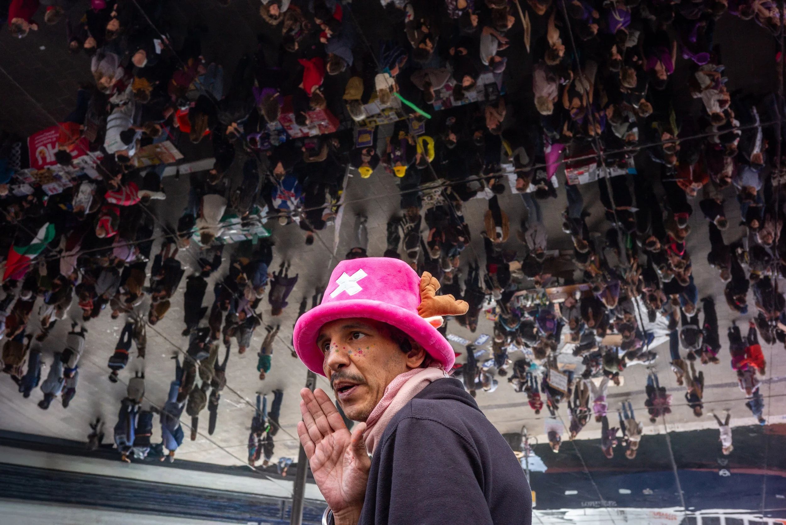 Une personne portant un chapeau rose en forme de star avec des motifs en forme de croix, faisant un geste avec la main et parlant. Au-dessus, on voit un grand miroir réfléchissant une foule de personnes dans une attitude diverse, dans un espace intér