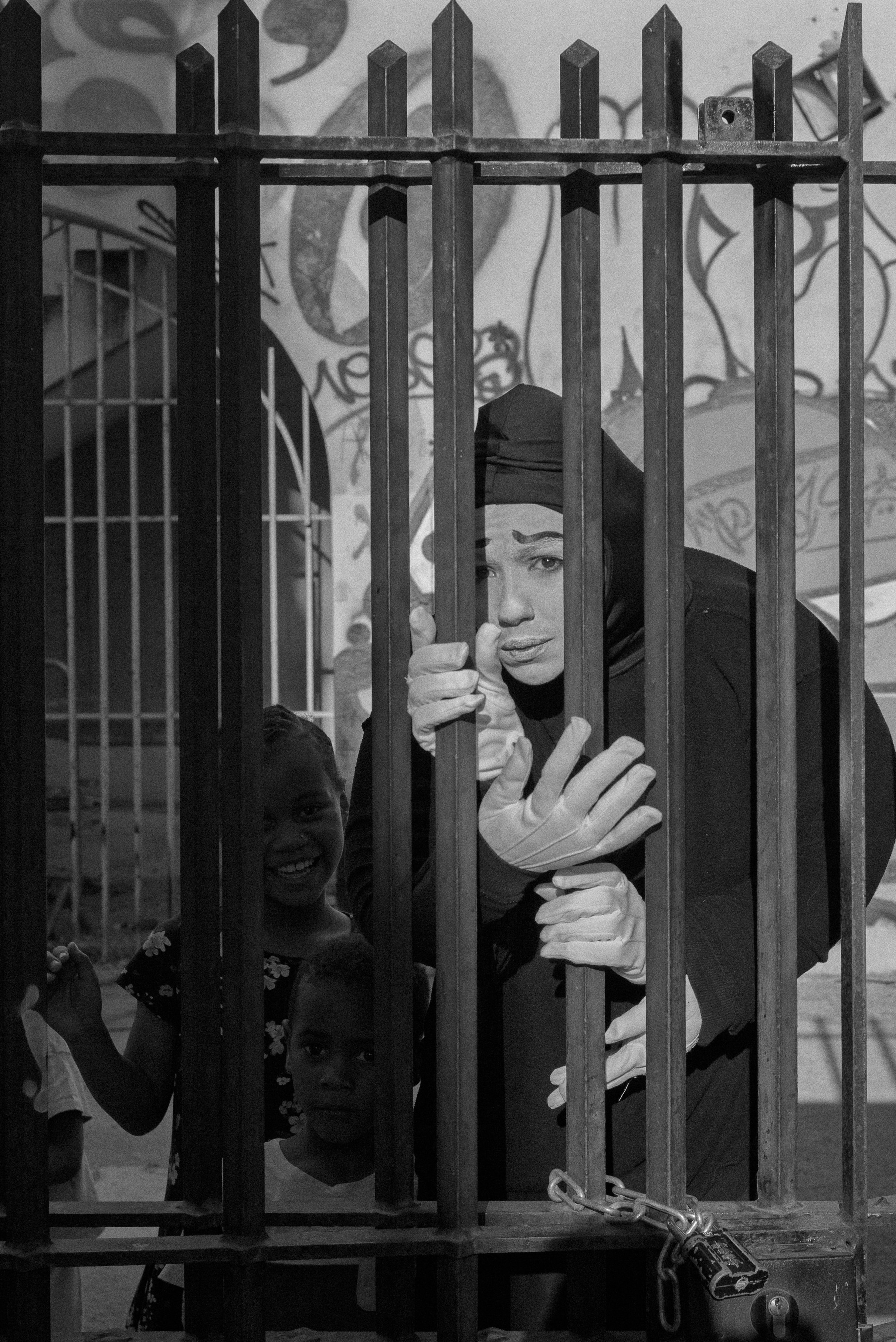 Une femme regarde à travers une grille de prison. Elle porte un hijab et des gants, et une clé est attachée à la serrure de la grille. Deux enfants sont visibles derrière la grille, dont une avec un sourire.