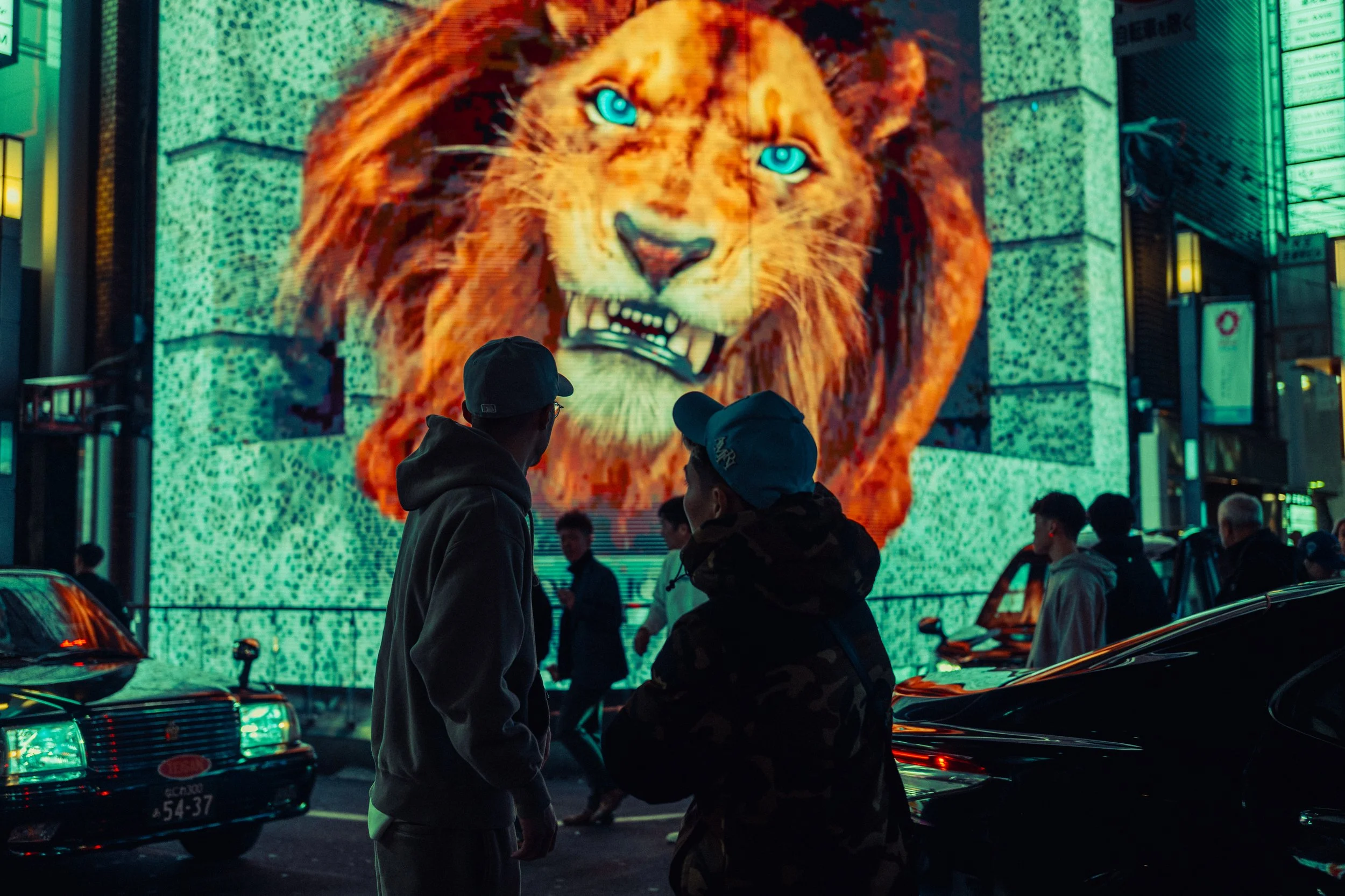 Deux jeunes hommes discutent devant un grand écran numérique affichant la tête d'un lion. La scène se déroule la nuit, avec des voitures en stationnement autour.