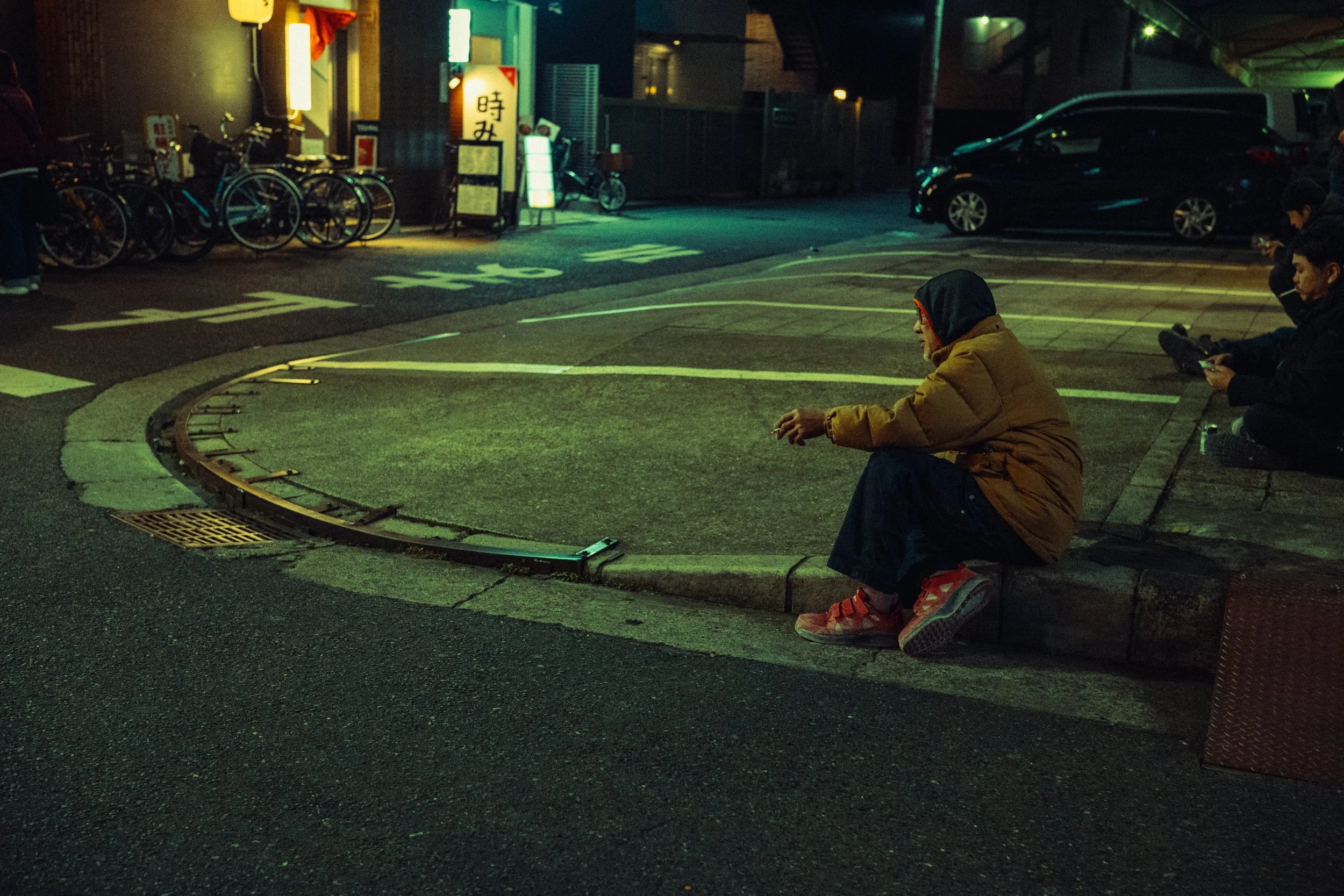 Une personne assise sur un bord de trottoir dans une rue éclairée par des lumières de nuit, portant une veste marron, des vêtements rouges et des chaussures de sport oranges.