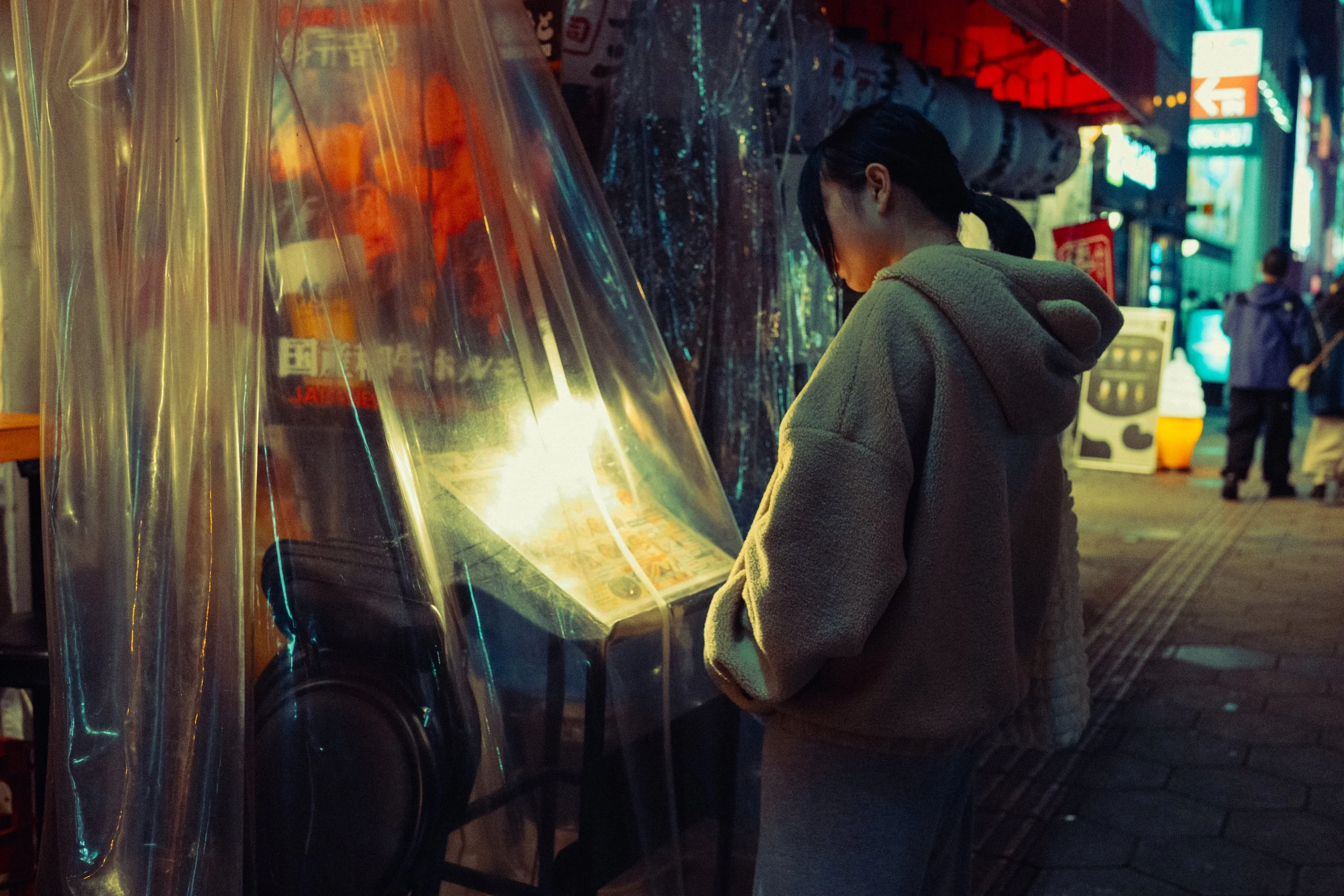Une jeune femme portant un manteau côtelé gris clair et une capuche regarde un stand de jeux ou de musique dans une rue éclairée la nuit, avec d'autres passants en arrière-plan.