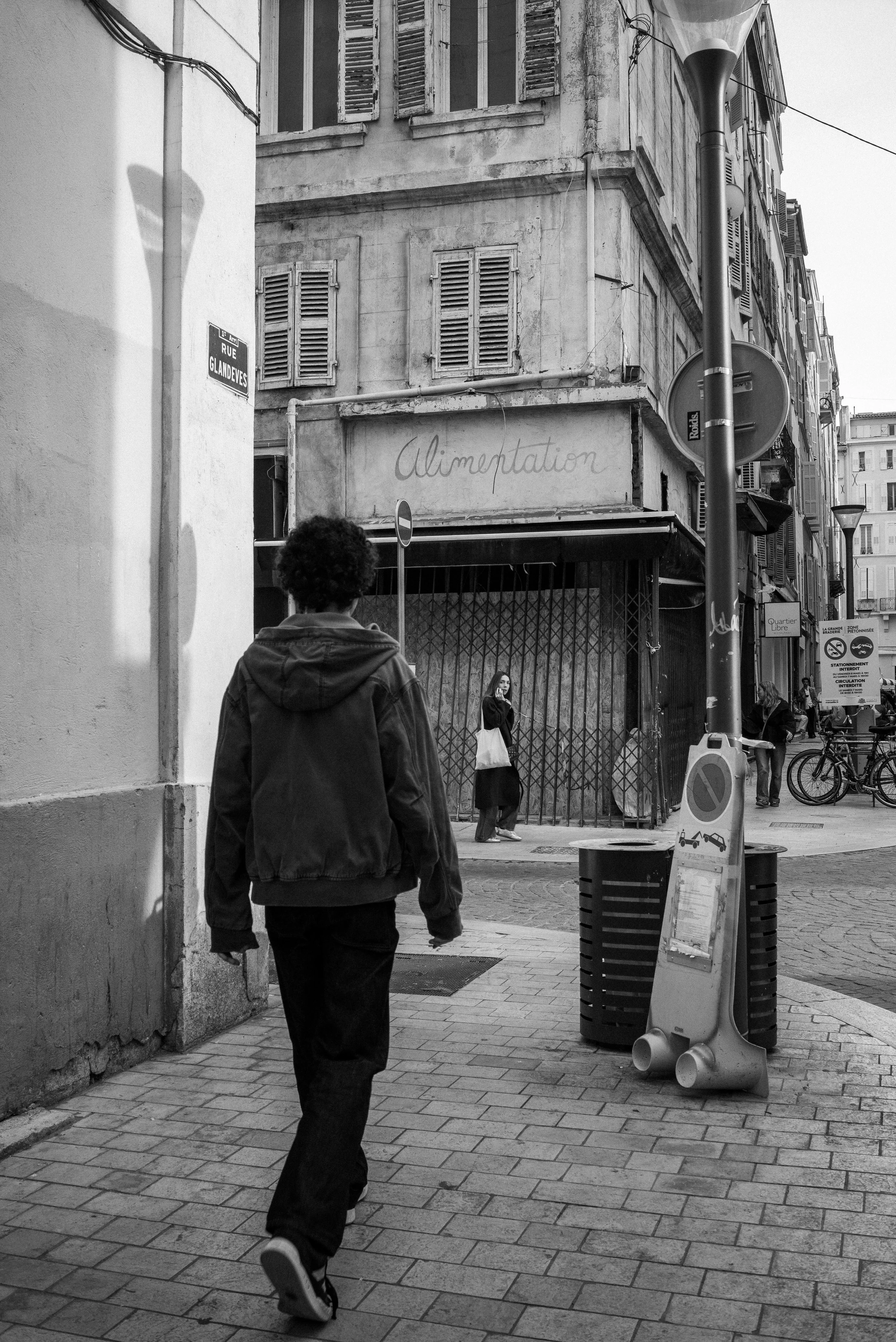 Une rue parisienne en noir et blanc avec un jeune homme marchant et une femme au téléphone devant un bâtiment avec un panneau "Alimentation".