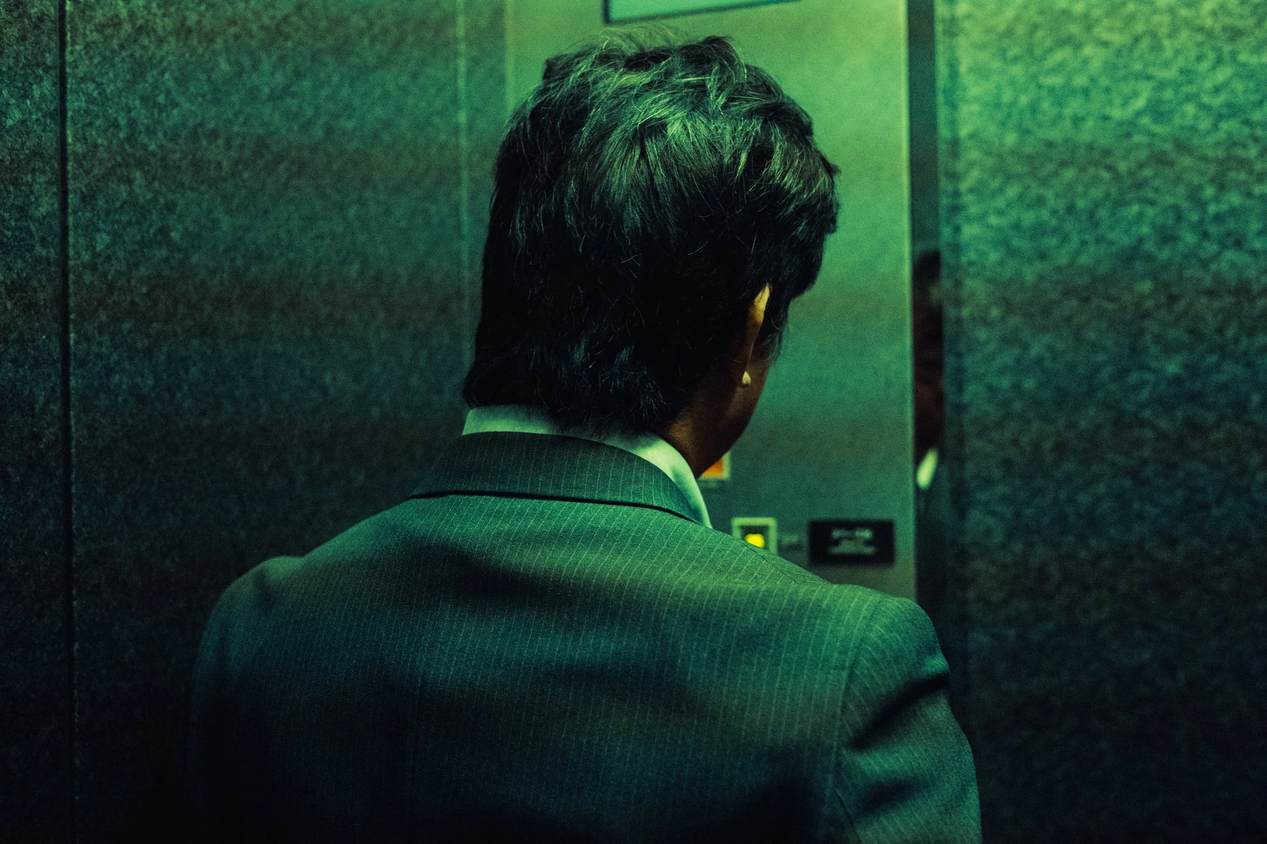 Un homme en costume regardant une fiche dans un ascenseur, vu de derrière, avec un éclairage vert.