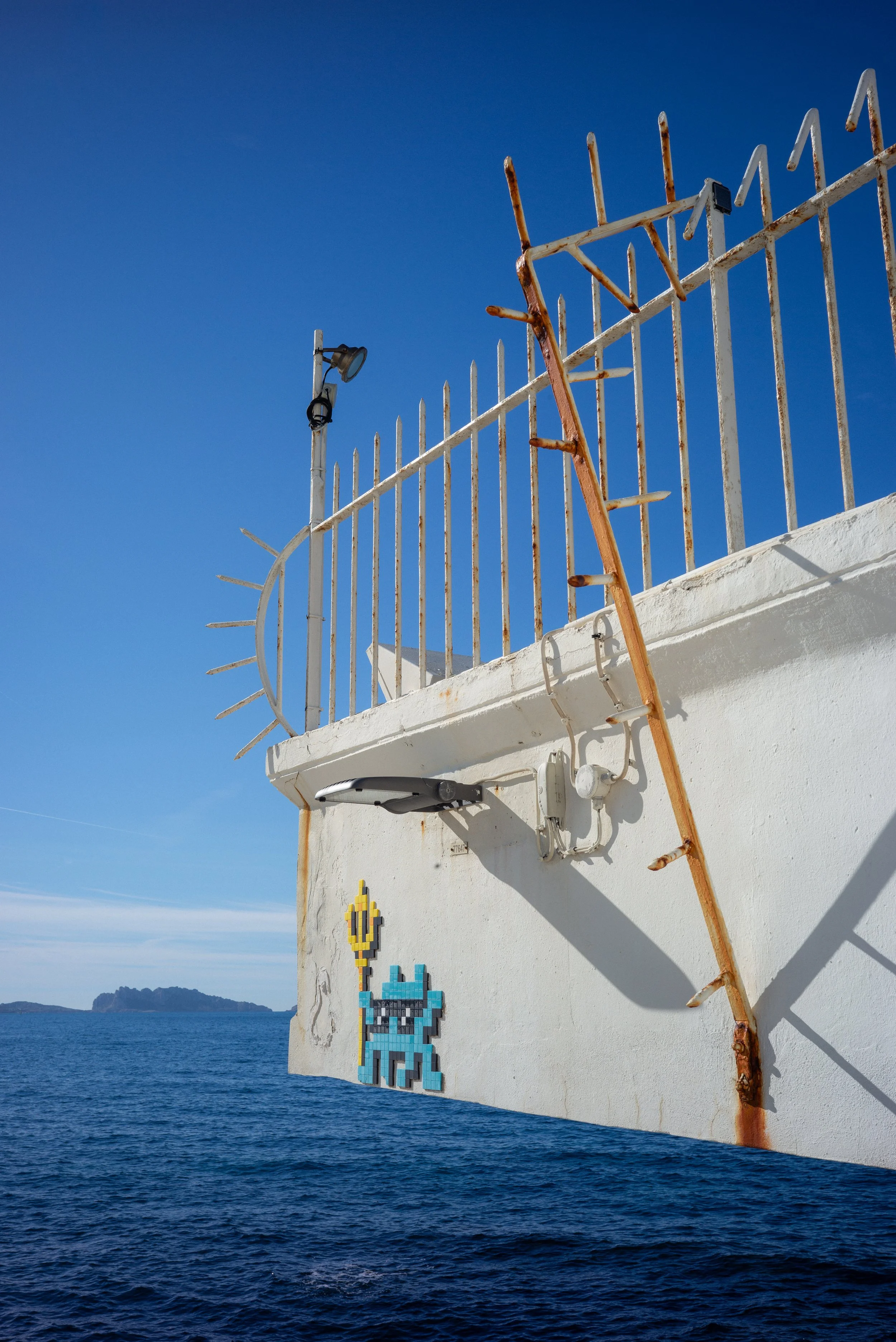 Vue de l'océan avec une structure de clôture en fer rouillé, un pixel art en mosaïque représentant un robot bleu, et un paysage d'île au loin sous un ciel clair.