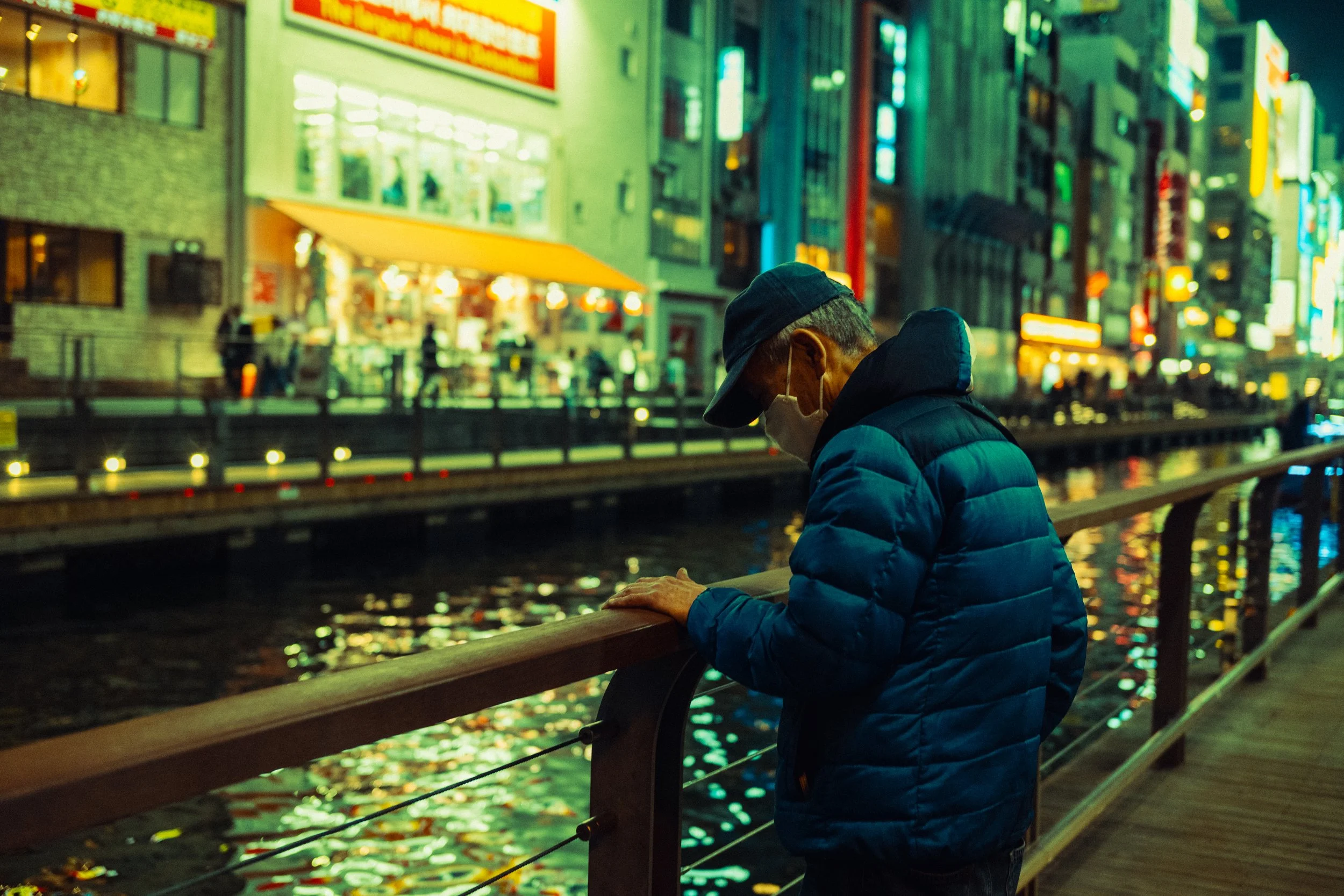Un homme porte un masque et une veste bleue, se tient près d'une rambarde au bord d'un canal ou rivière en ville la nuit, avec des lumières colorées et des enseignes lumineuses en arrière-plan.
