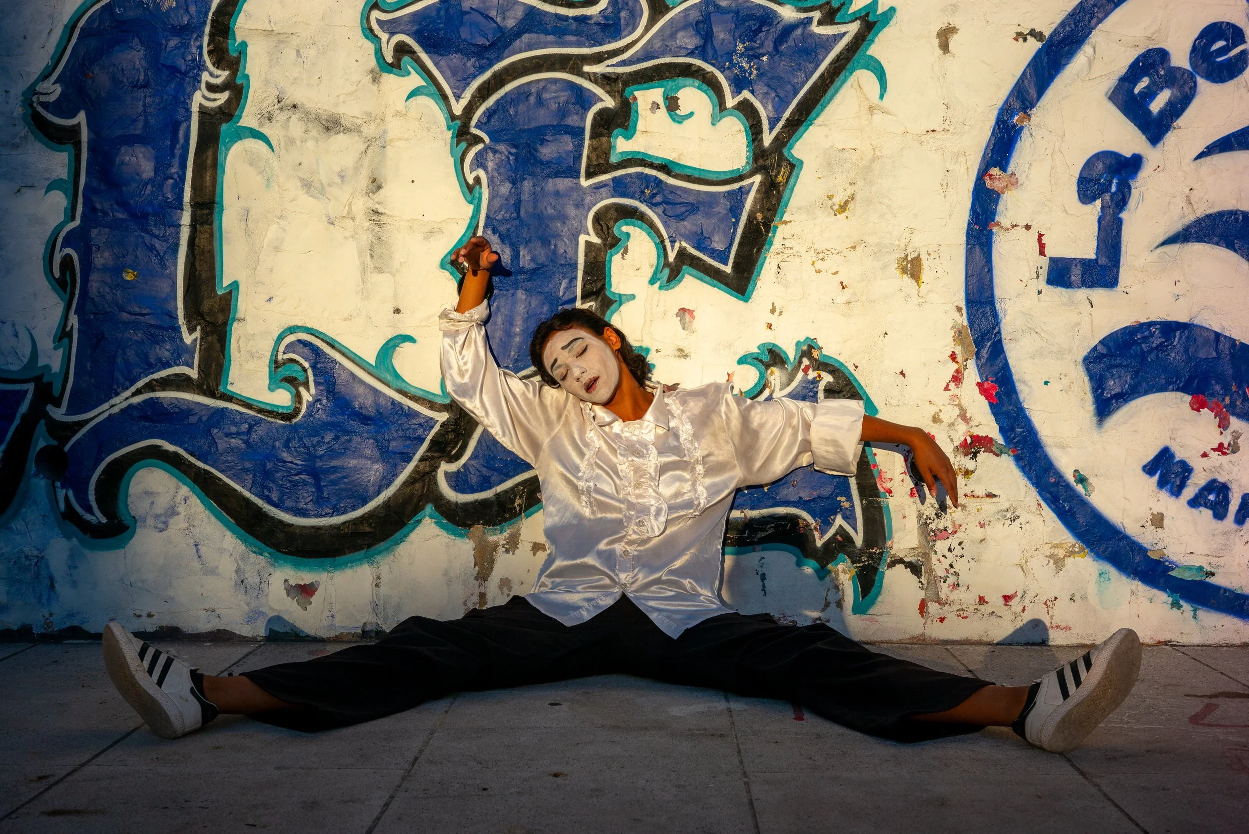 Une femme danseou pose en extéieur devant un mur couvert de graffiti, portant une chemise blanche, des pantalons noirs, et des chaussettes à rayures.