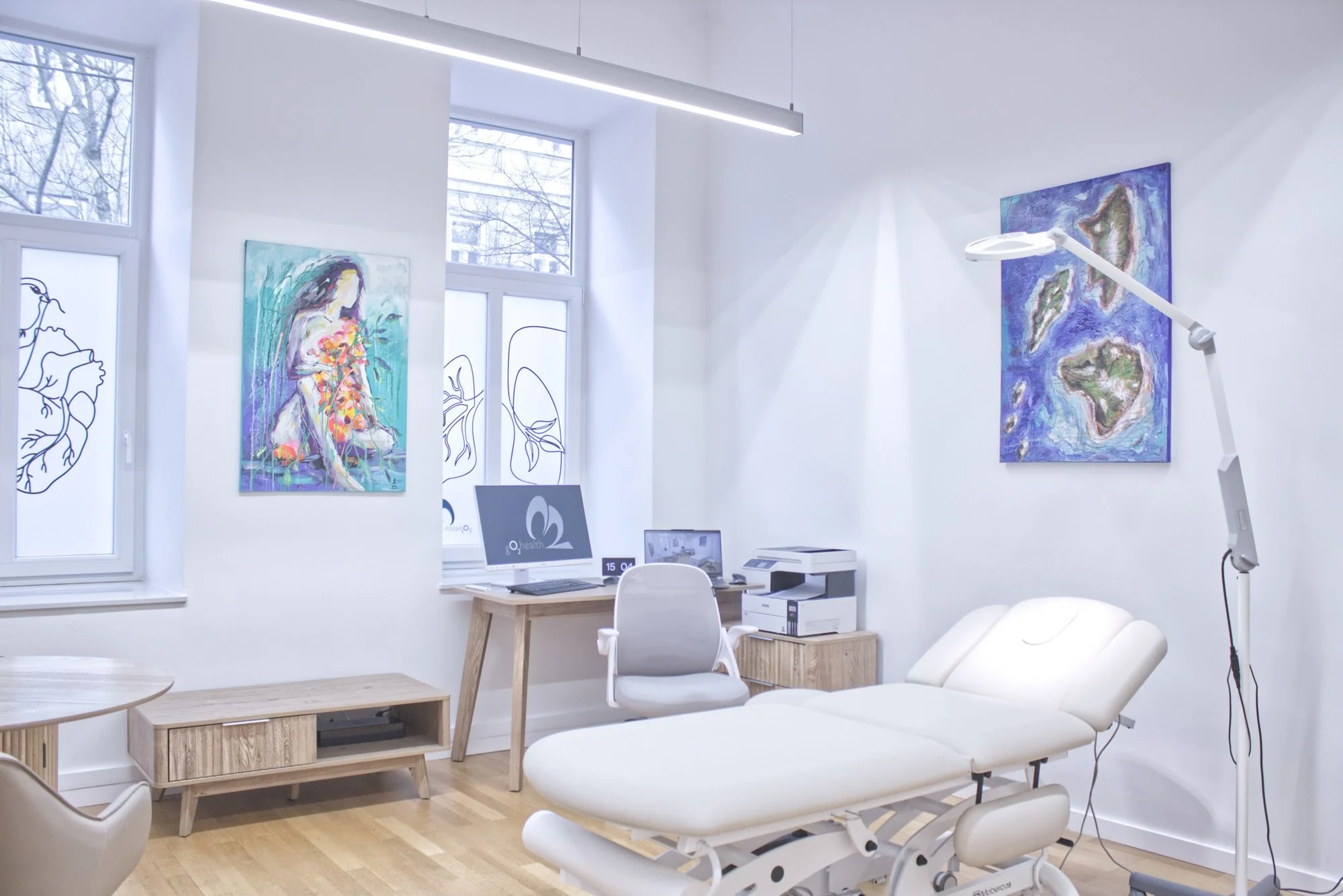 Ein Behandlungszimmer in einer Arztpraxis mit weißen Wänden, mehreren modernen Kunstwerken, einem weißen Behandlungstisch, einem Schreibtisch mit Computer und Drucker, einem weißen Stuhl und einer operativen Lampe.