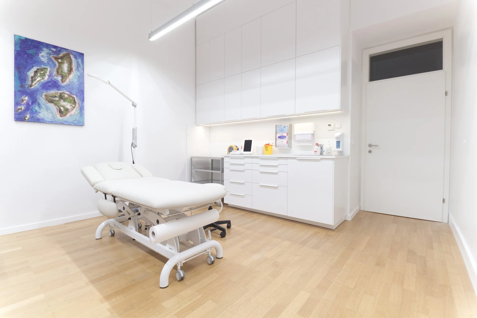 Ein weißer Behandlungsstuhl in einem medizinischen Behandlungsraum, an der Wand befindet sich ein Kunstwerk mit blauen und grünen Inseln. Der Raum ist modern gestaltet mit weißen Schränken und verschiedenen medizinischen Utensilien auf der Ablage.