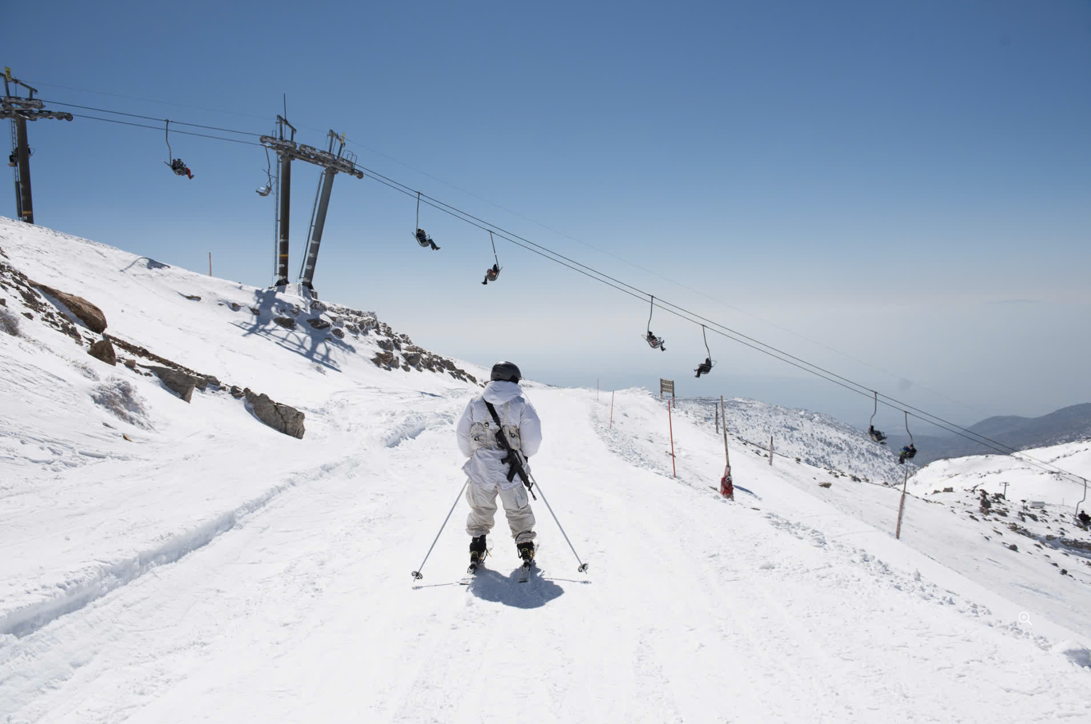A snowy ski resort with skiers and snowboarders riding the ski lift on a clear day. A skier is in the foreground wearing white gear, facing away from the camera, while others are visible on the lift in the background.