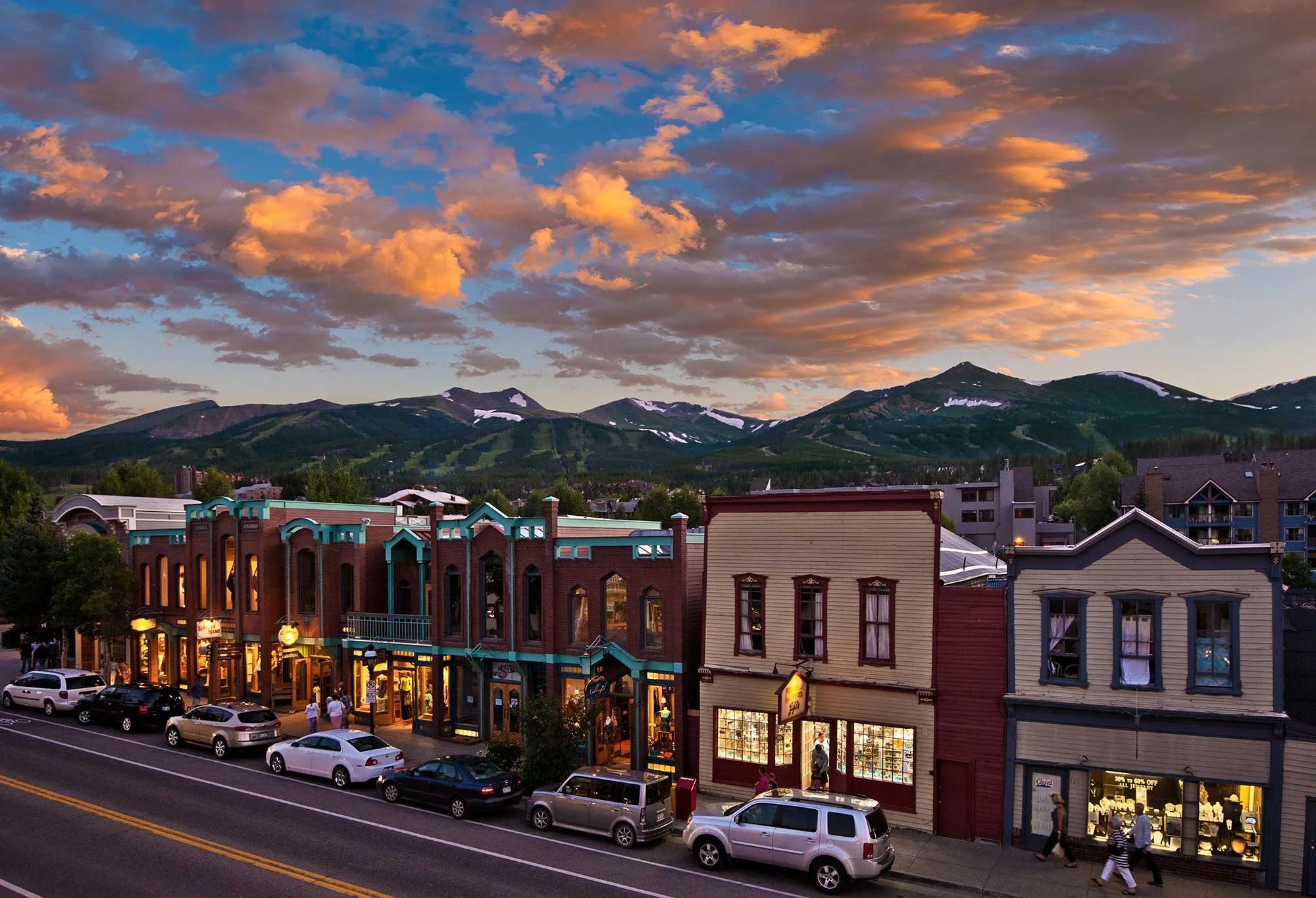 Town street with parked cars, storefronts, hills with snow patches, and a colorful sky during sunset.
