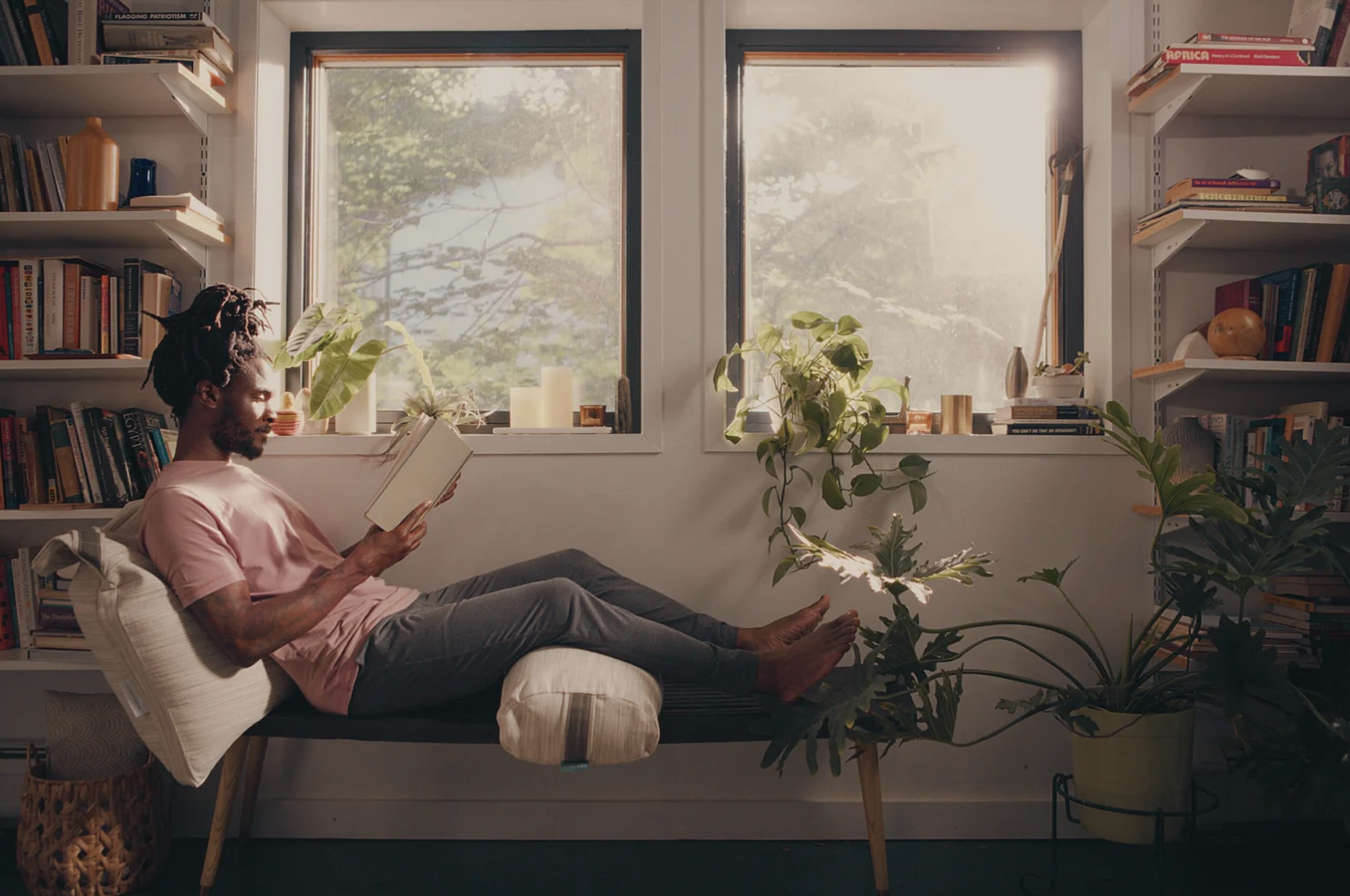 Man with dreadlocks reading a book while lying on a padded bench in front of a window with sunlight, surrounded by houseplants and bookshelves.