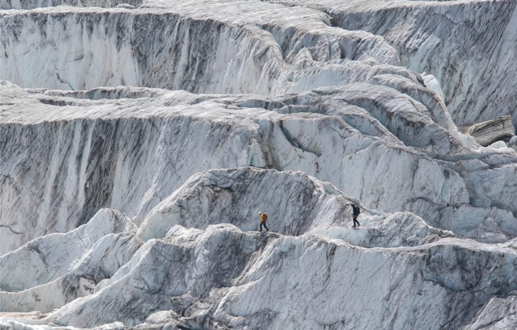 Two hikers walking across a vast glacier with blue and white ice formations.