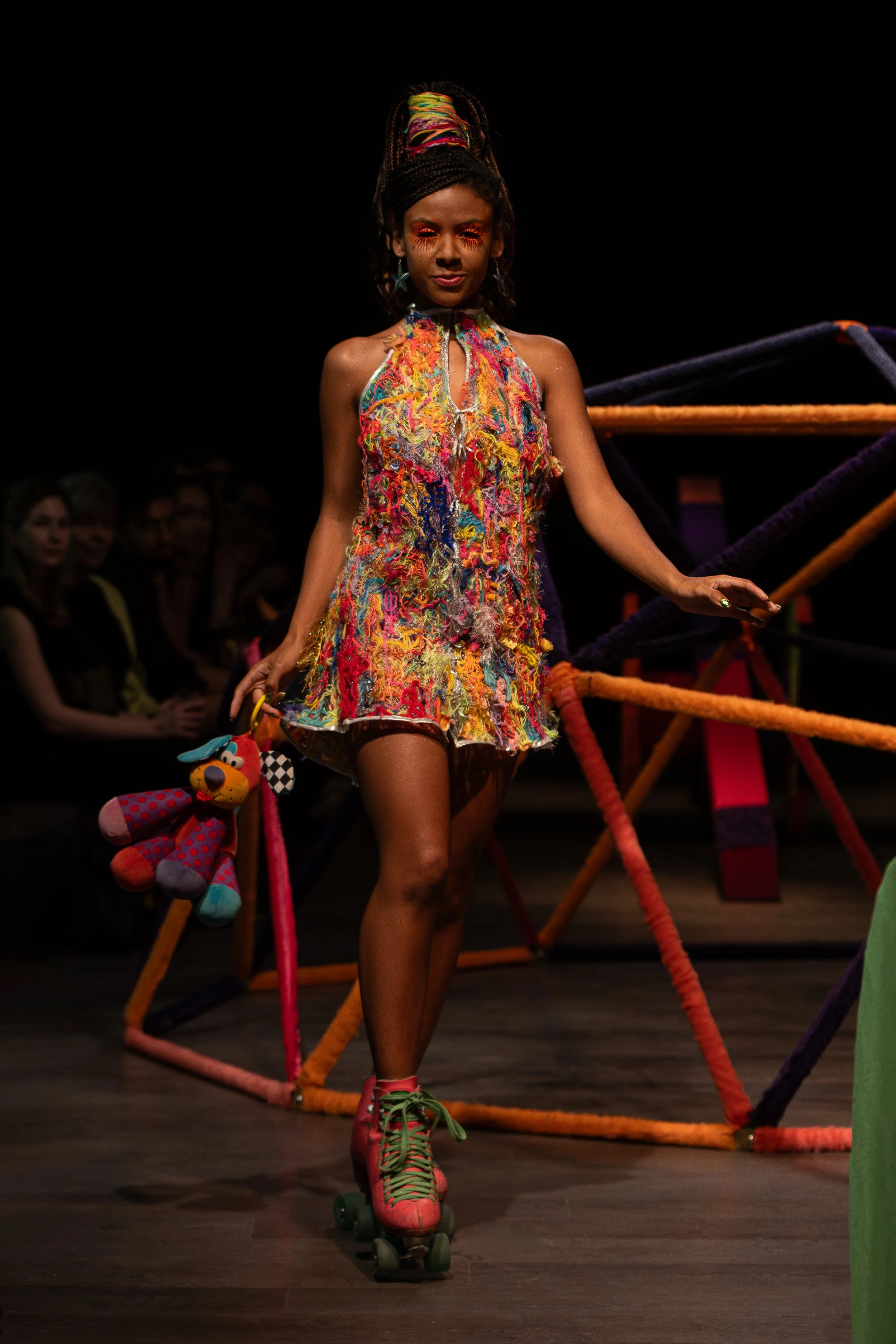 A person with dreadlocks, wearing colorful makeup, a vibrant sleeveless dress, and roller skates, walking on a stage with a dark background and an audience visible in the background.