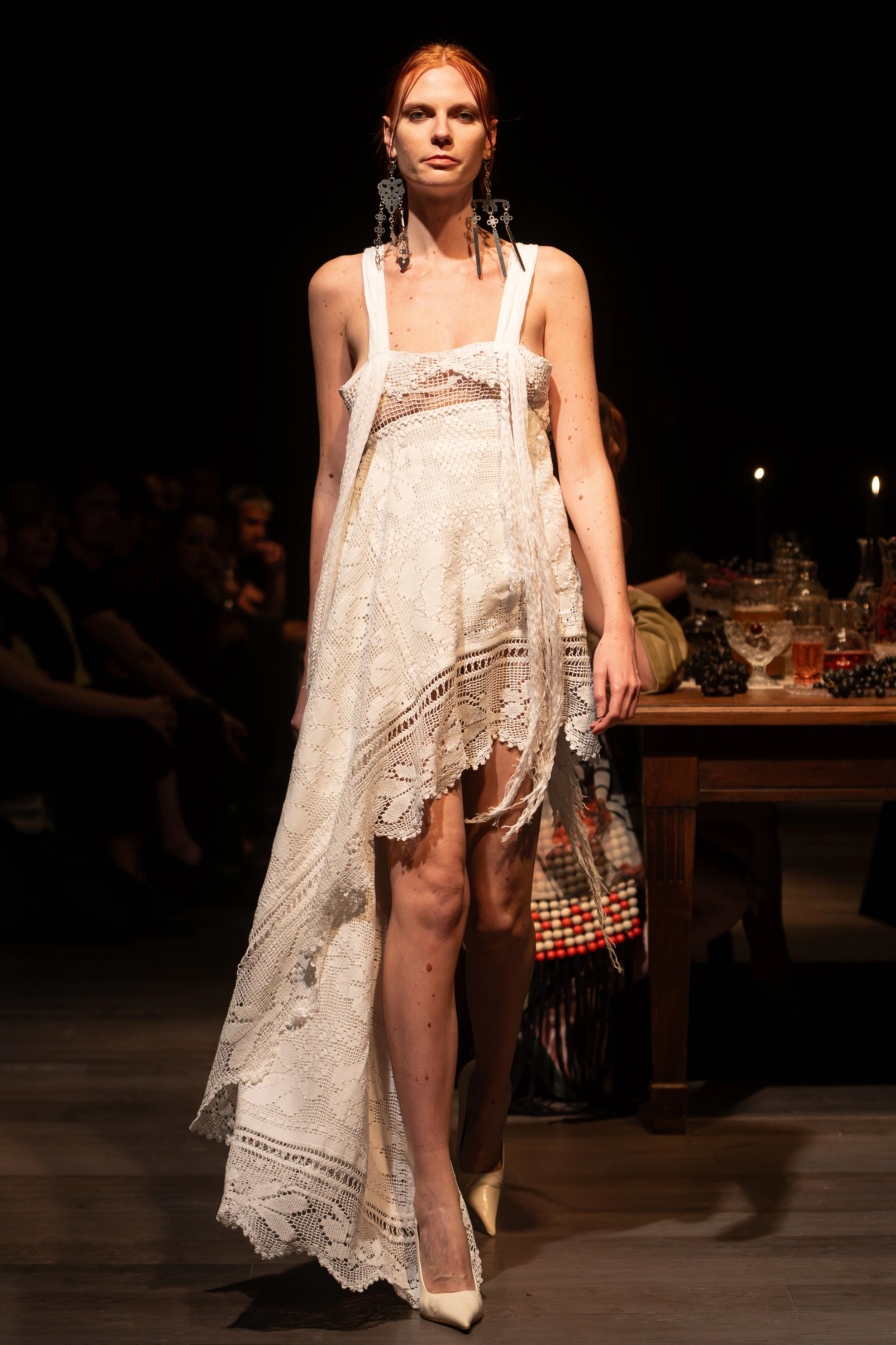 A woman with red hair wearing a white lace dress and earrings, walking on a runway during a fashion show.