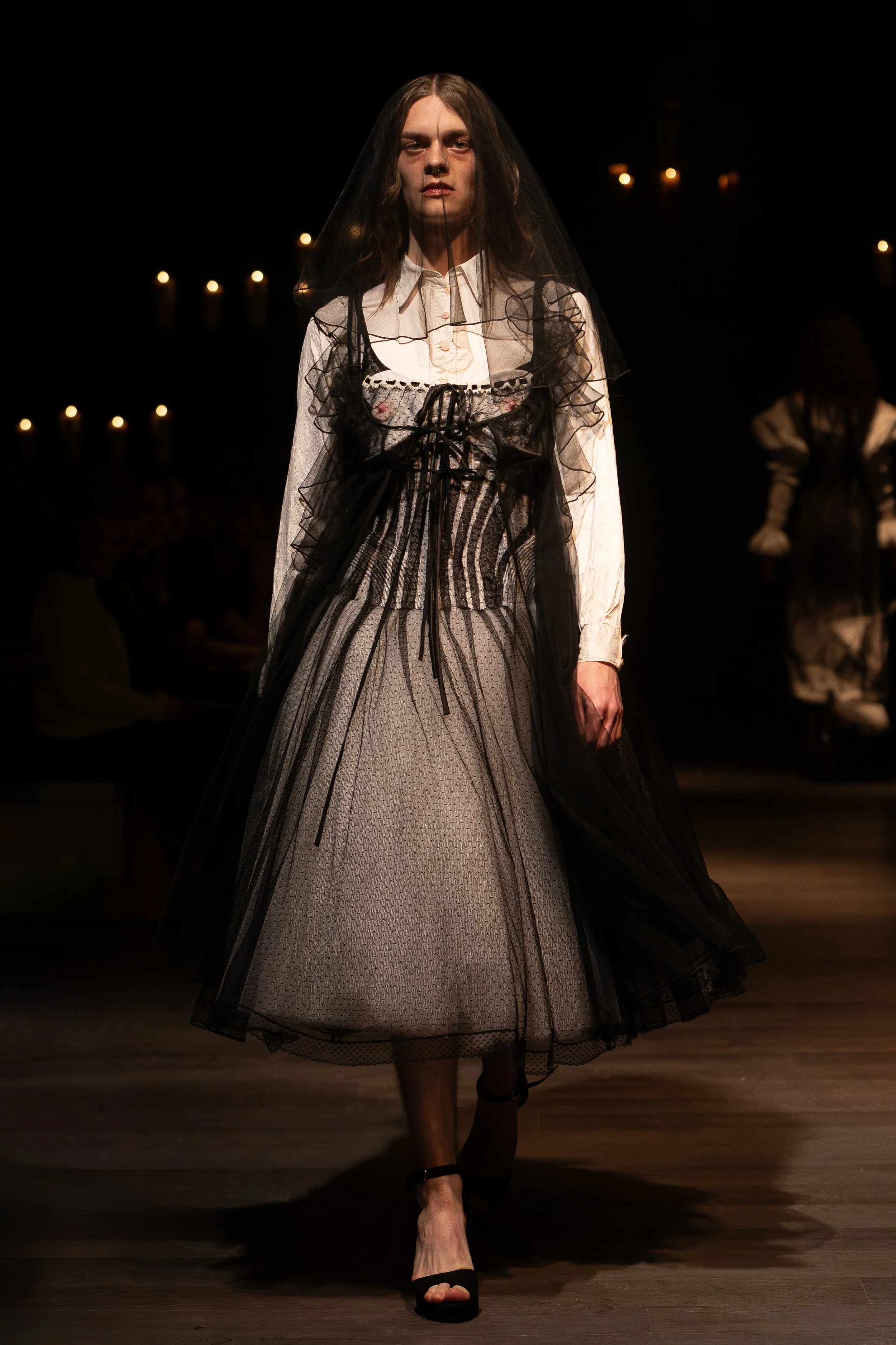 Model walking down a runway wearing a vintage-inspired dress with a layered tulle skirt, long sleeves, and a high neckline, with a sheer black veil covering most of her face, in a dark setting with candles in the background.