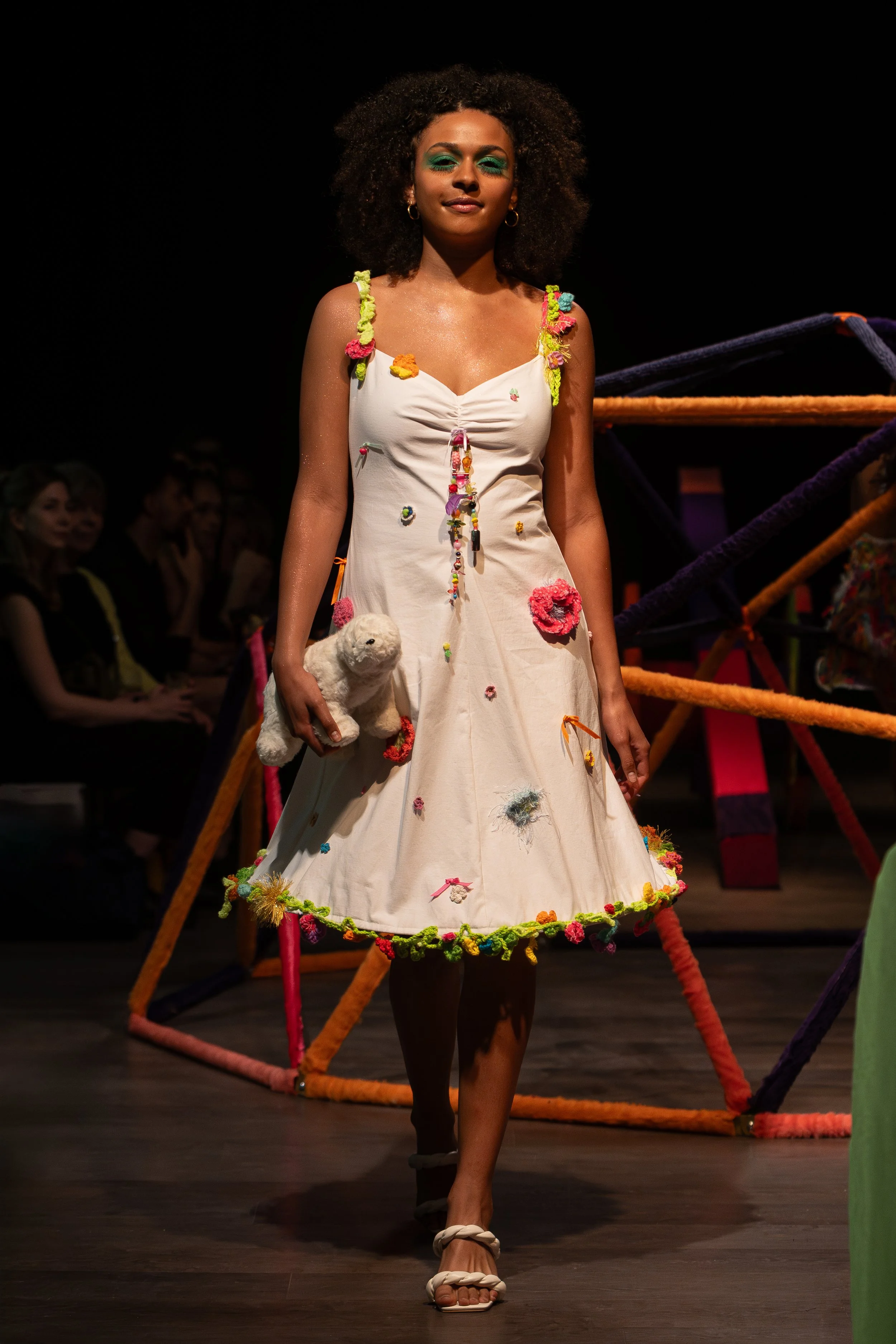 A woman walking on a runway wearing a white dress decorated with colorful flowers and holding a plush toy, with a dark background and an audience in the distance.