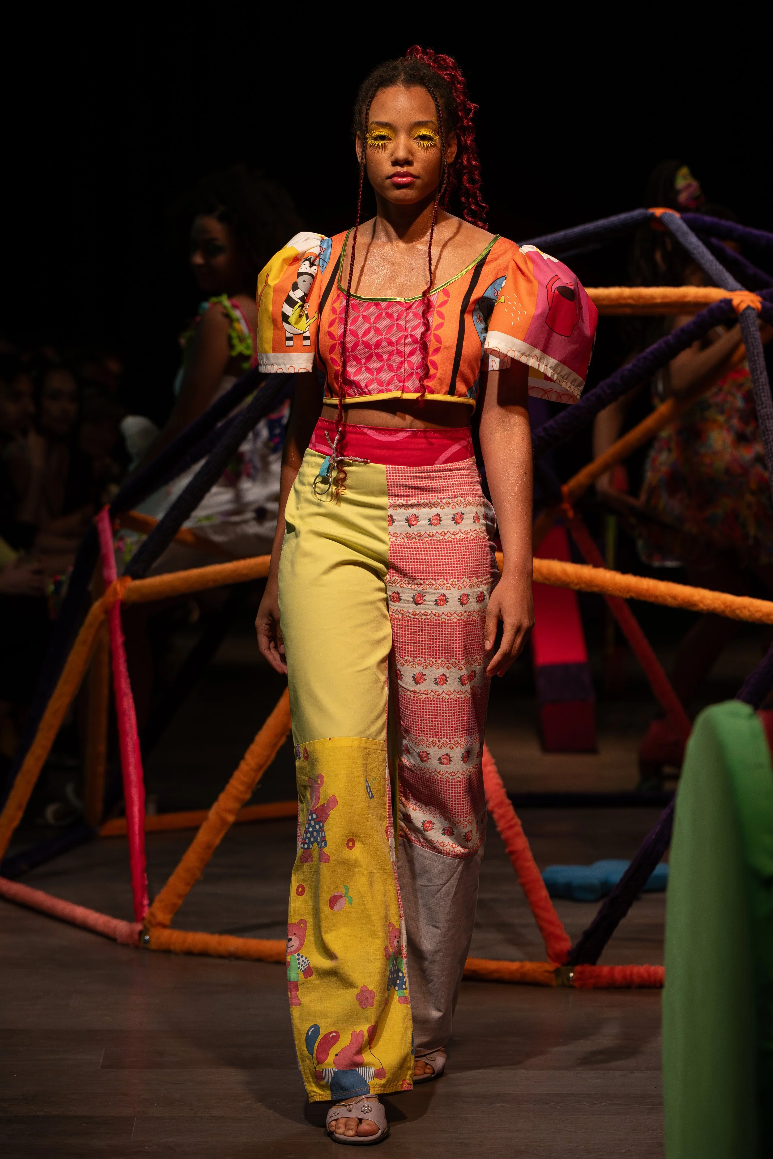 Young woman walking on stage during a fashion show, wearing colorful patchwork pants with cartoon prints, a vibrant patchwork top, with colorful yellow eyeshadow and styled curly hair, with a dark background and other models in colorful outfits behin