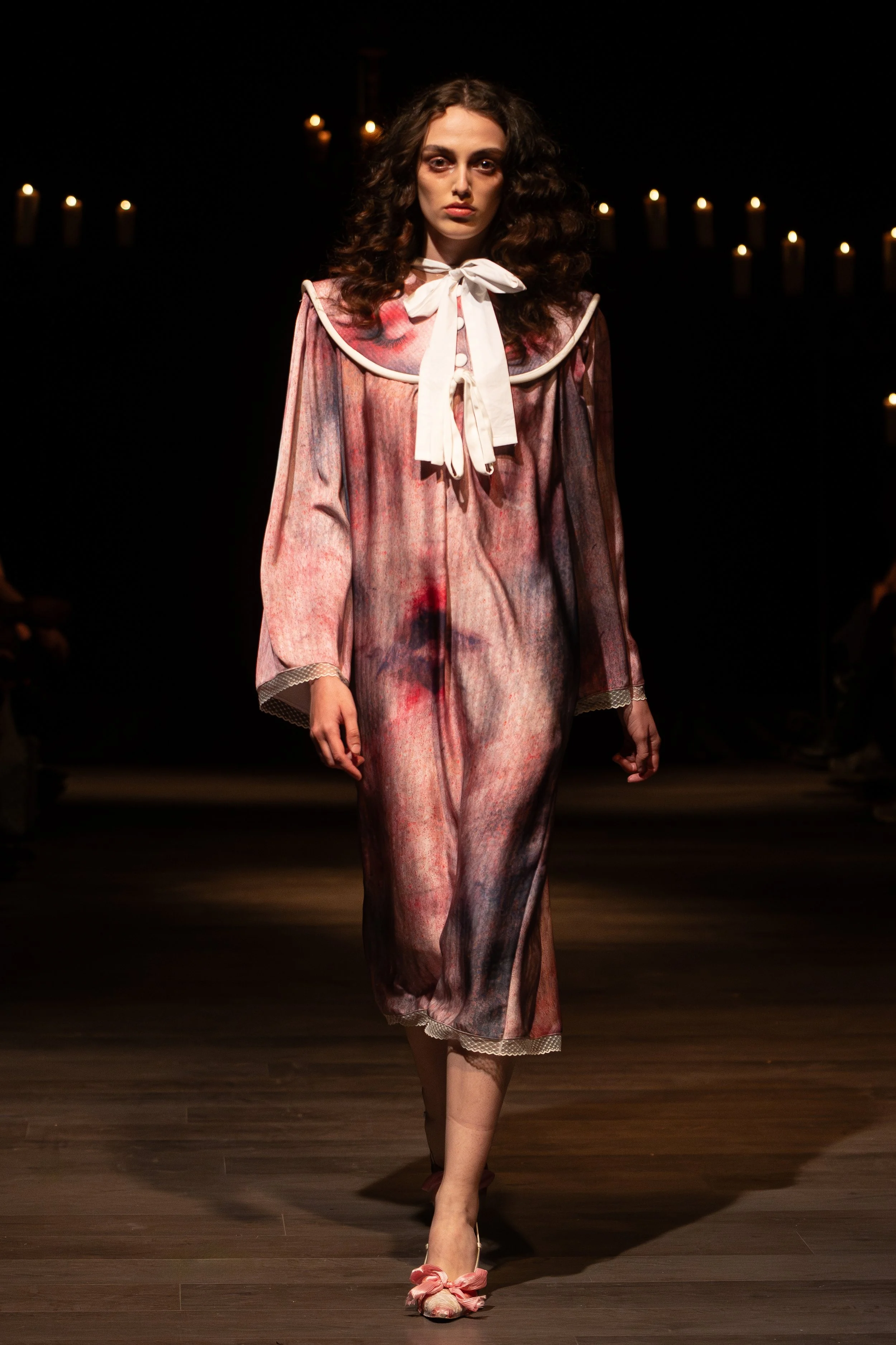 A female model walking down a runway in a vintage-style, tie-dye pink and purple dress with a white bow tie at the collar. The background is dark with yellowish candles on the wall, creating a moody atmosphere.
