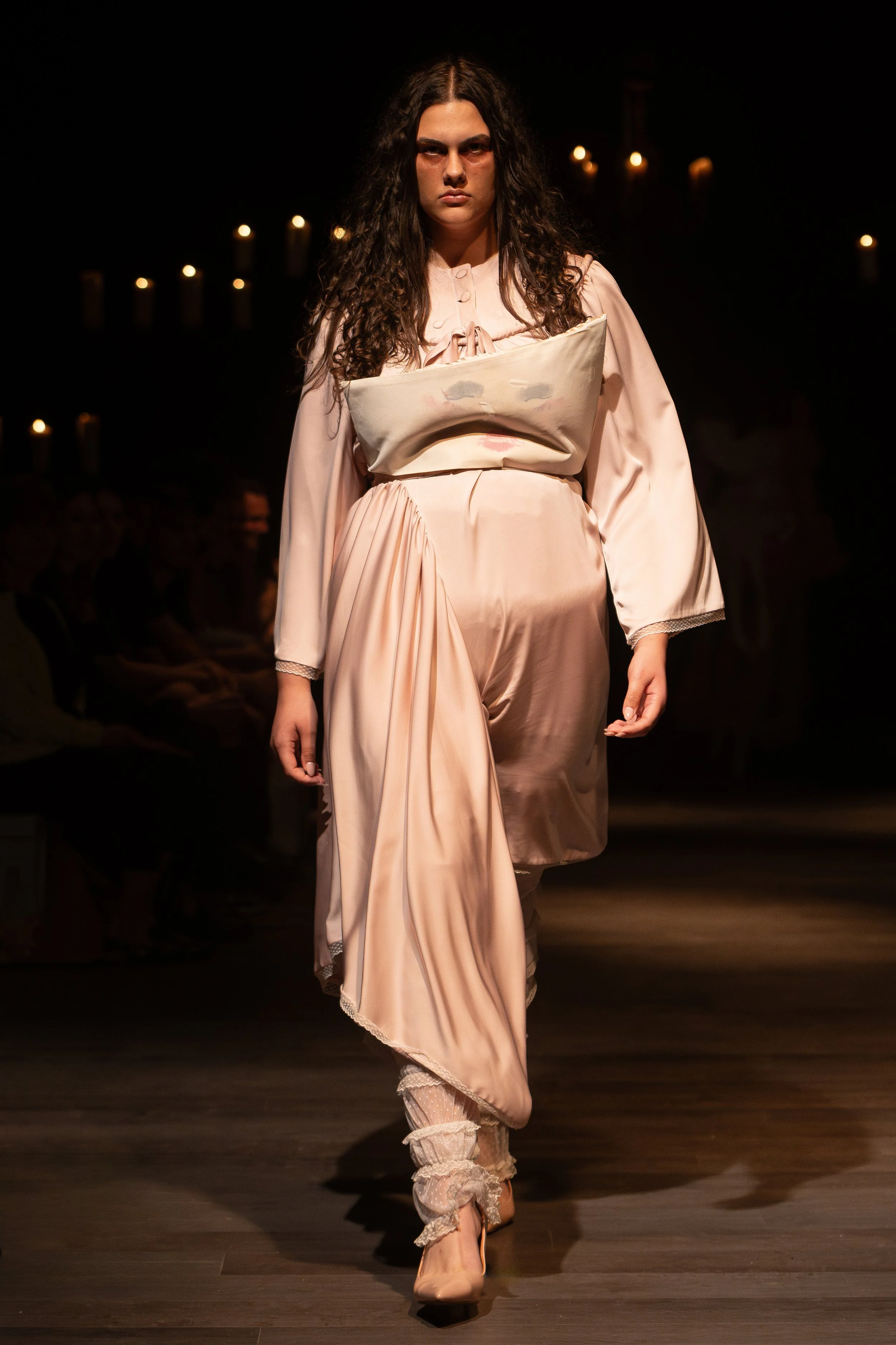 A woman with long curly hair walking down a runway, wearing a vintage style satin pink dress with lace details, a large satin bow on her chest, and lace-up shoes with bows, during a fashion show with candles in the background.