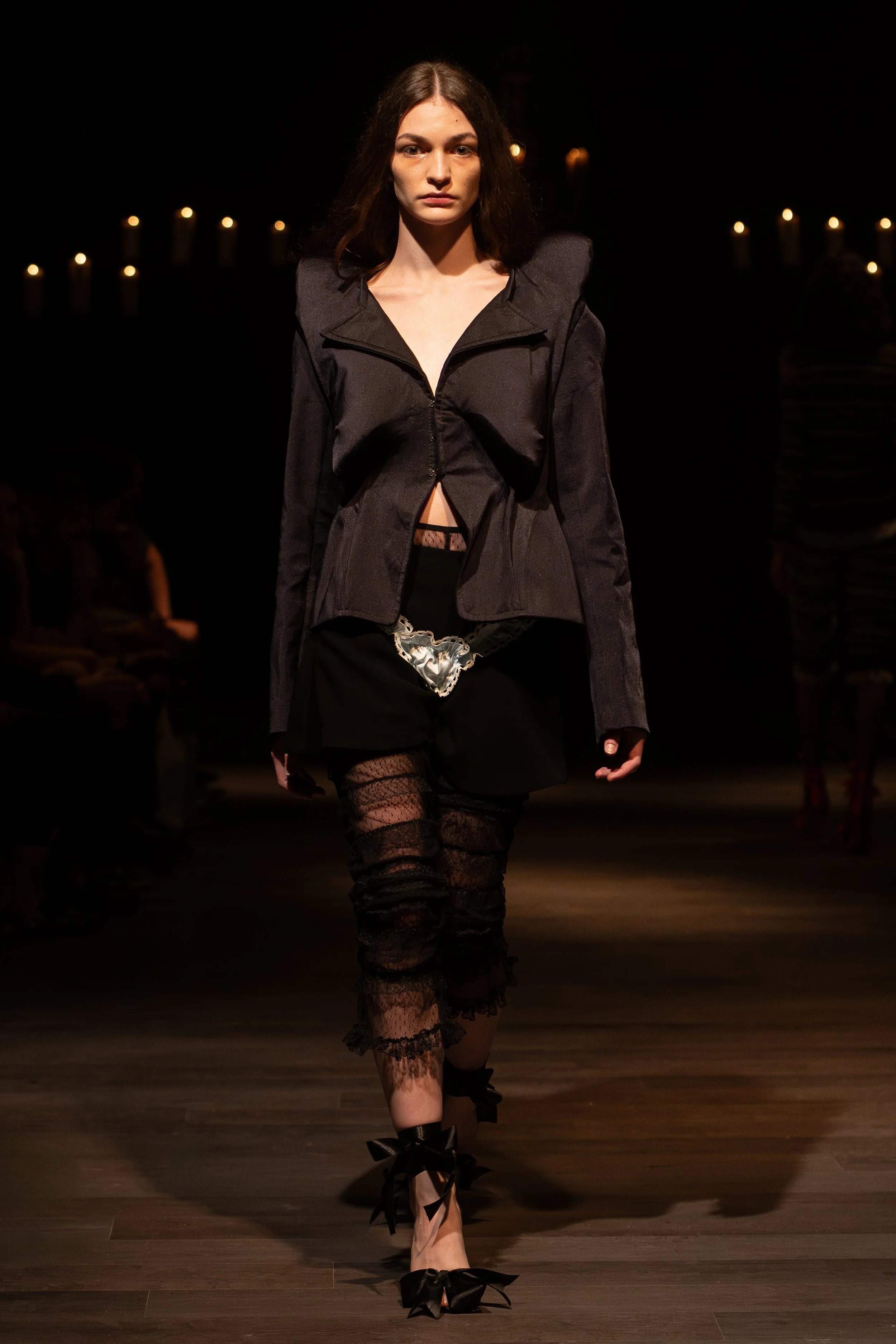 A female model is walking down a runway in a fashion show, wearing a dark jacket, lace stockings, and high-heeled shoes with bows at the ankle.
